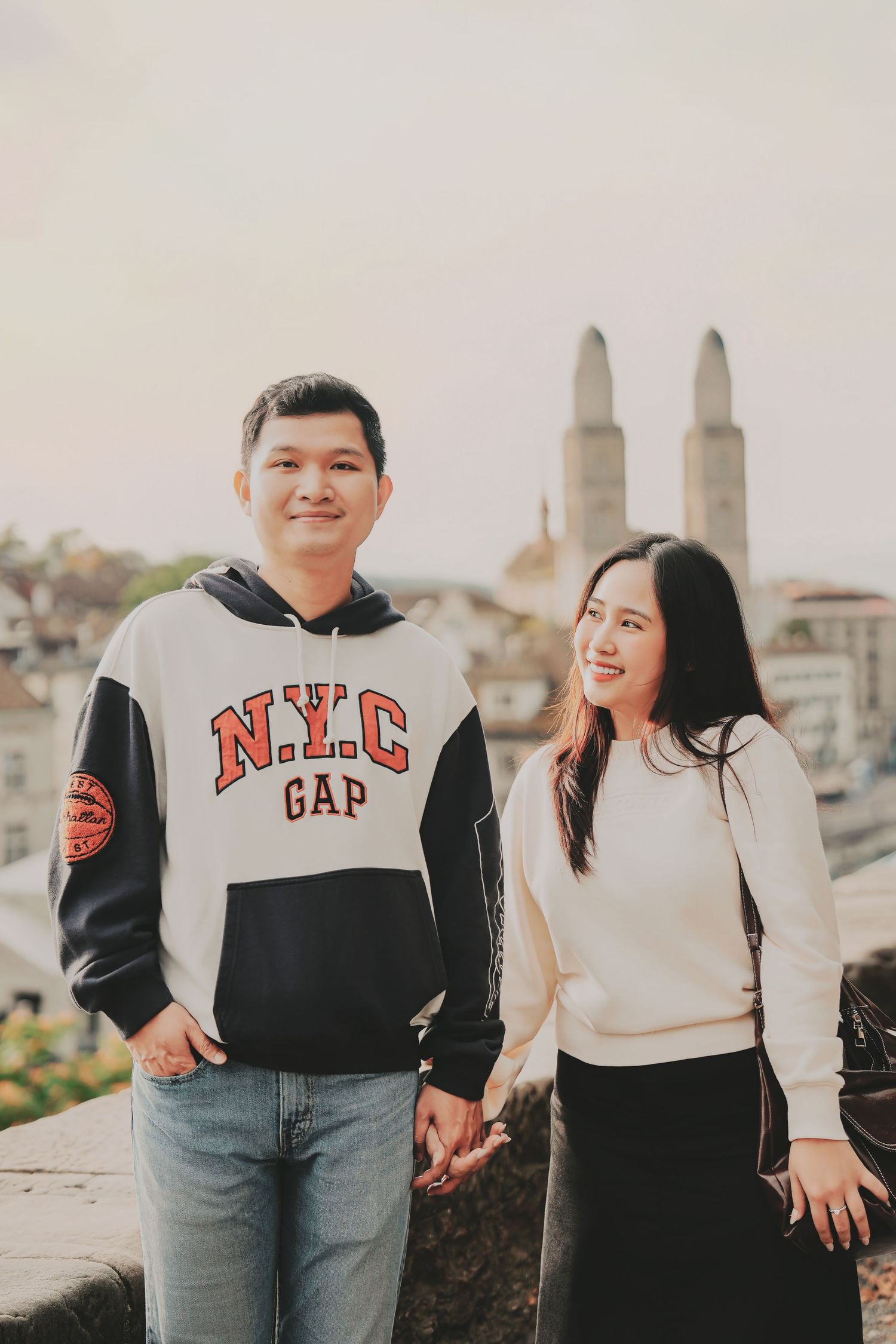 Young Asian couple smiling and holding hands with Zurich old town and twin towers in background.