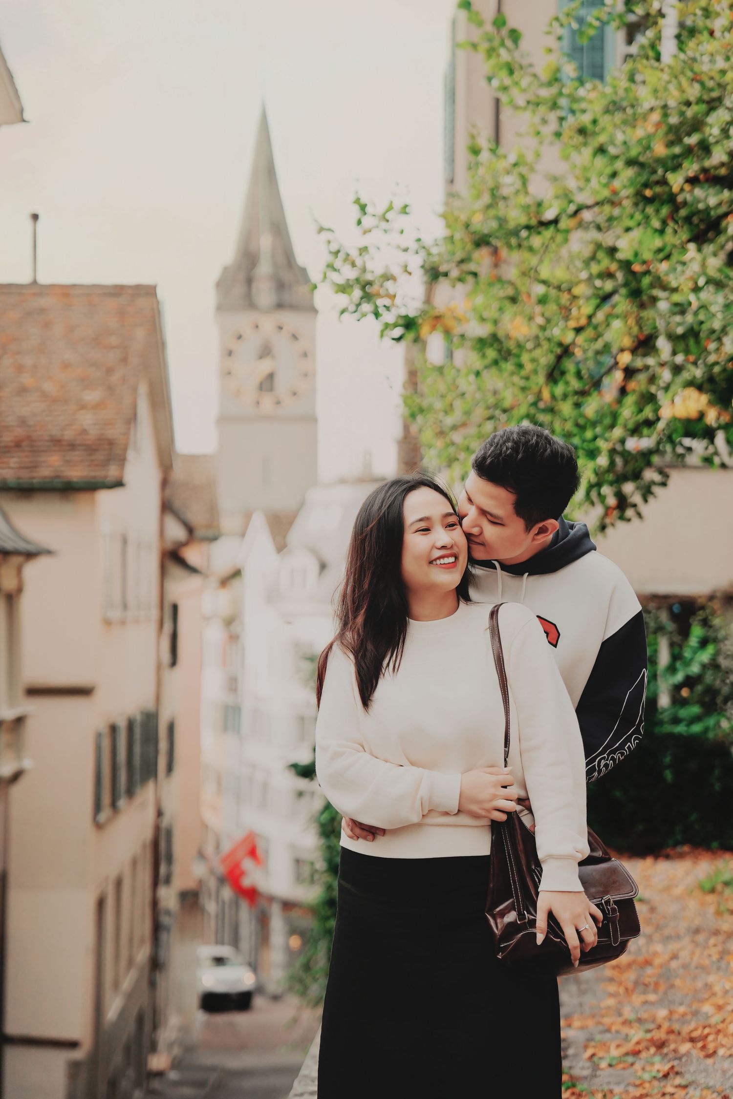 Man kissing woman's cheek as she smiles on a charming Zurich old town street with Swiss architecture.