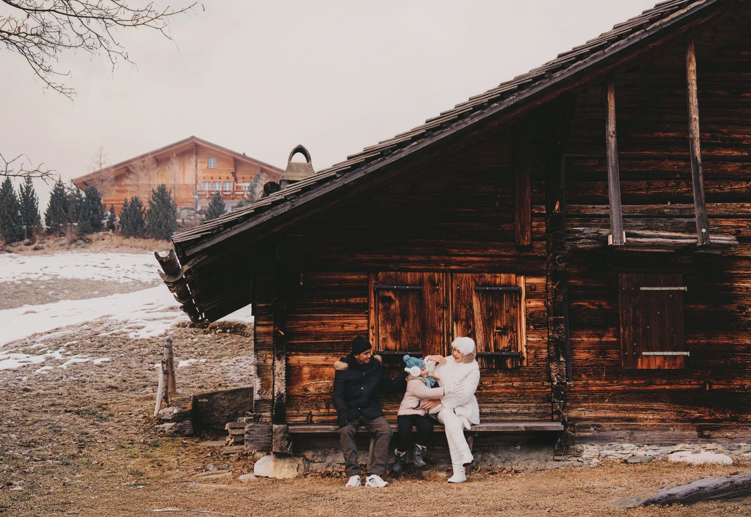 Family of three relaxing outside rustic wooden alpine cabin with snowy mountain scenery behind.