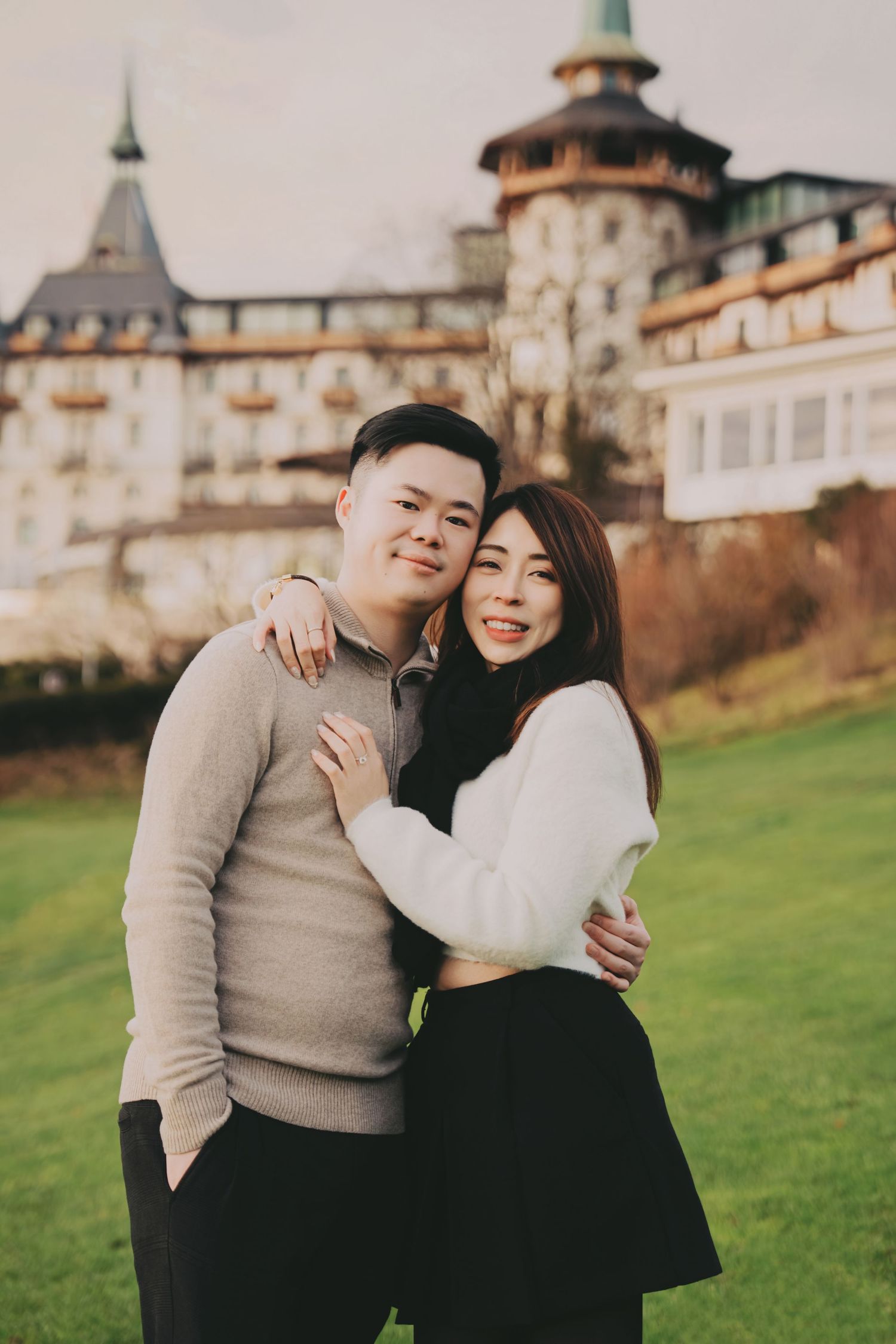 Smiling couple posing together on green grass with a grand European castle-style hotel behind them.
