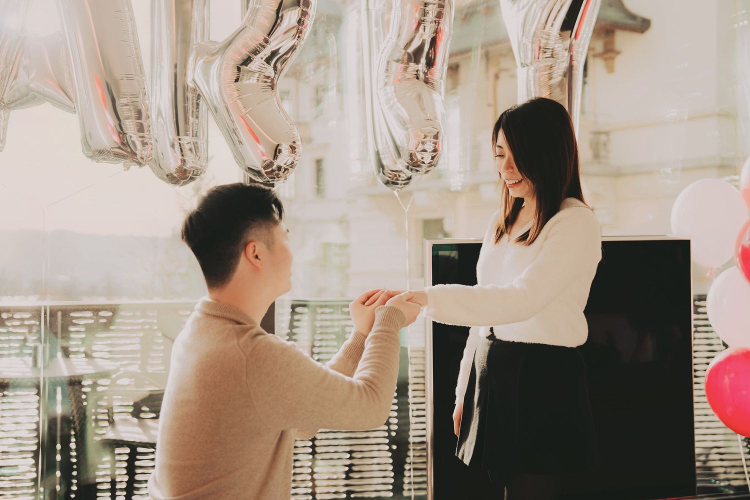 Man on one knee proposing to woman with silver letter balloons and pink balloons in decorated room.