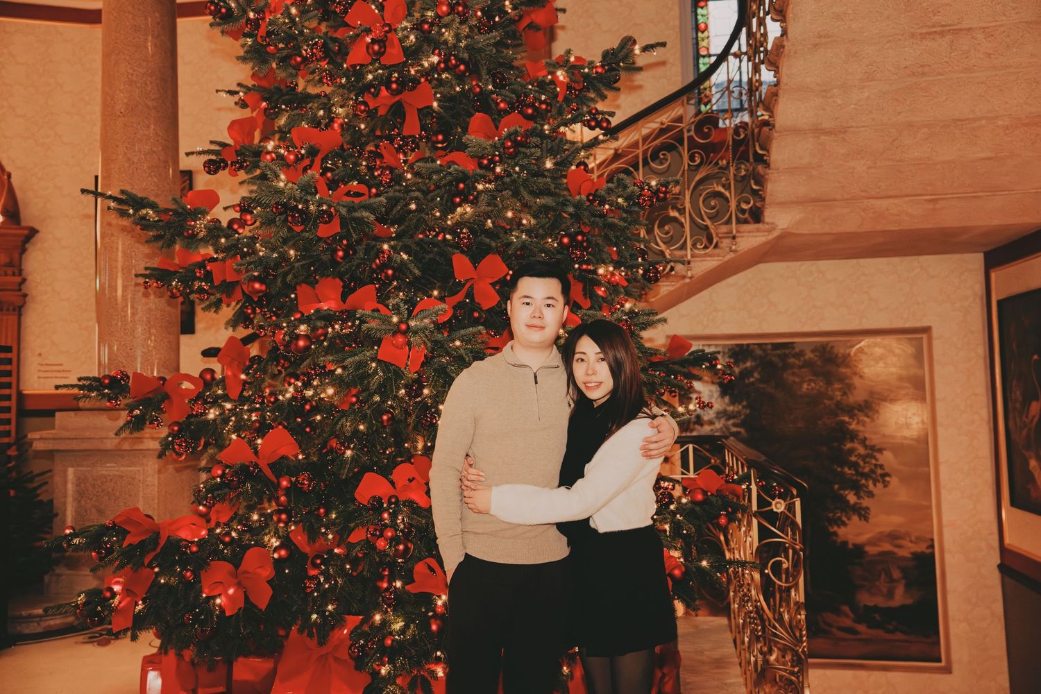 Couple embracing in front of large decorated Christmas tree in elegant holiday setting.
