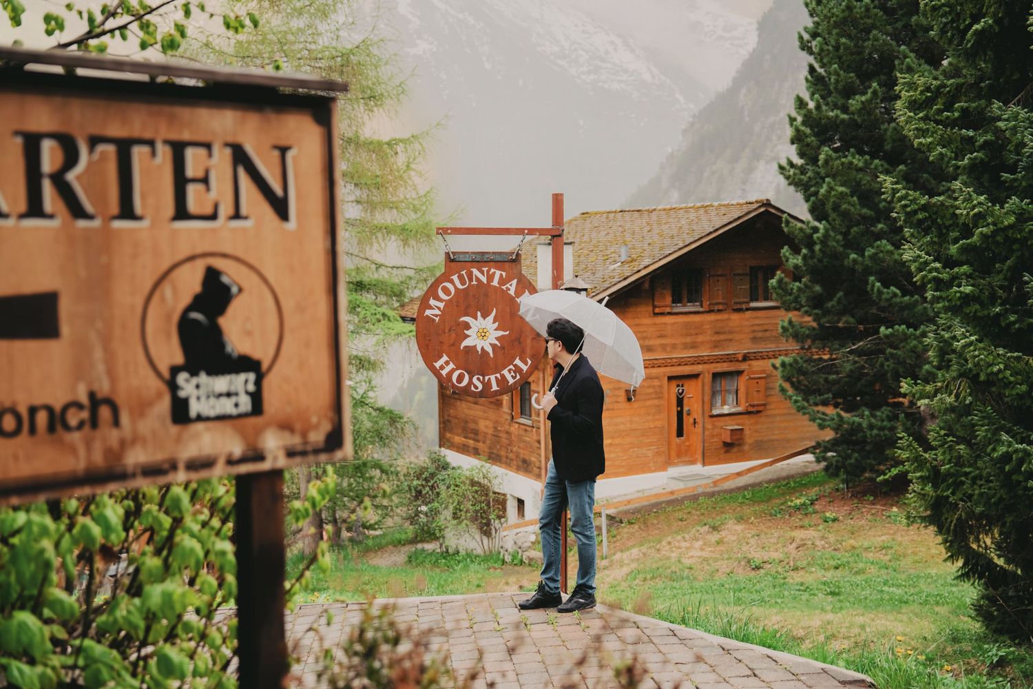 Person standing with white umbrella near Mountain Hostel wooden chalet in rainy alpine village.