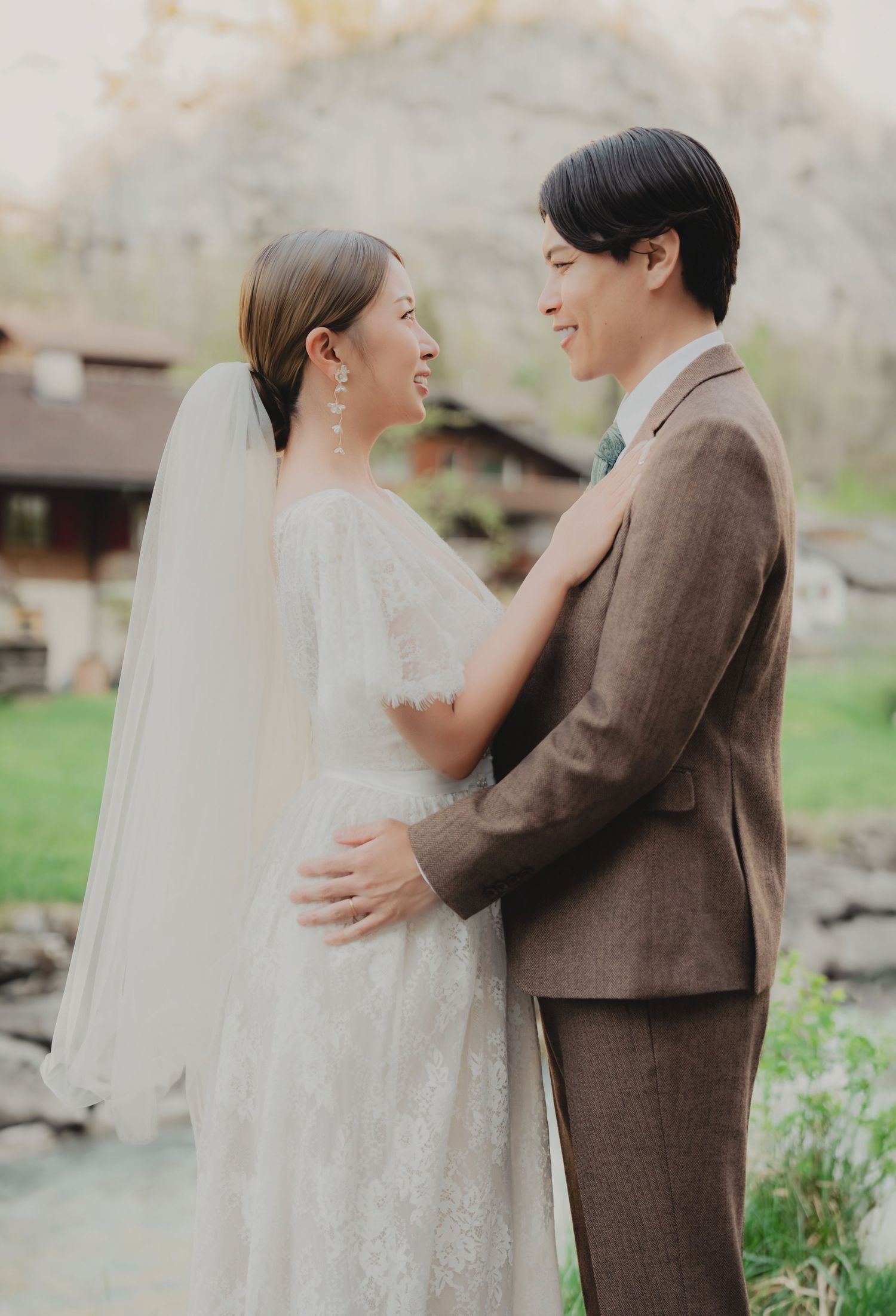 Couple in wedding attire share a tender moment, bride in lace gown and groom in brown suit by a stream.