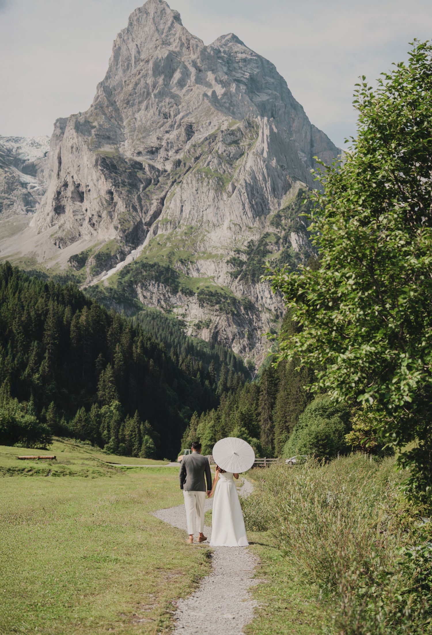 Wedding couple strolling on a narrow mountain trail with a white parasol against a towering rocky peak.