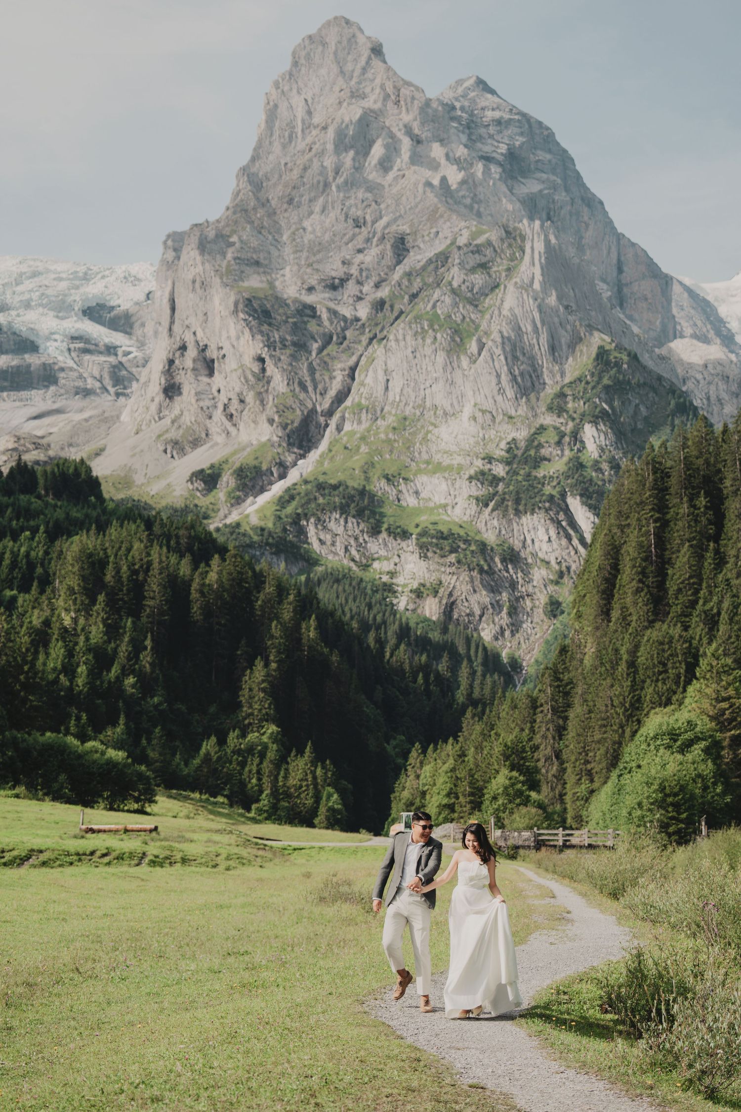 Bride and groom walk a mountain path with a dramatic rocky alpine peak towering above green forest behind them.