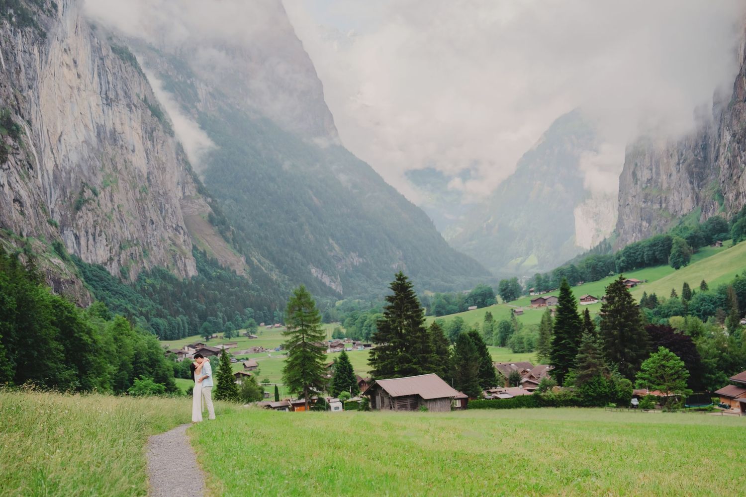Couple standing on a path in a vast green Swiss valley surrounded by towering misty mountains.