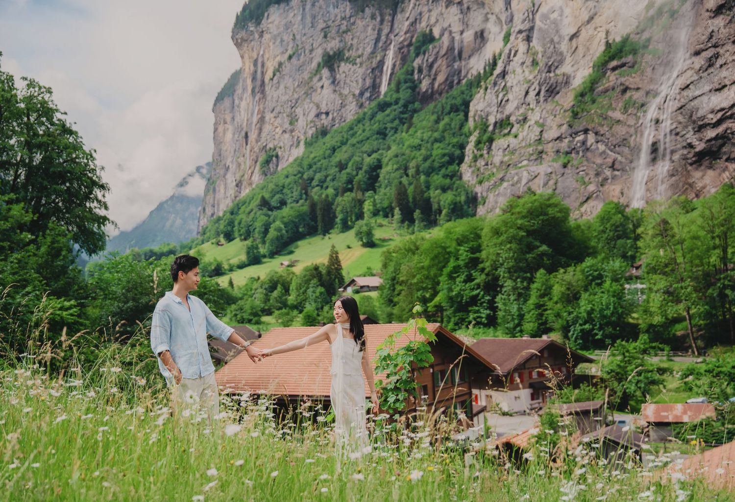 Couple holding hands in a lush green meadow with a waterfall and dramatic cliffs in Lauterbrunnen, Switzerland.