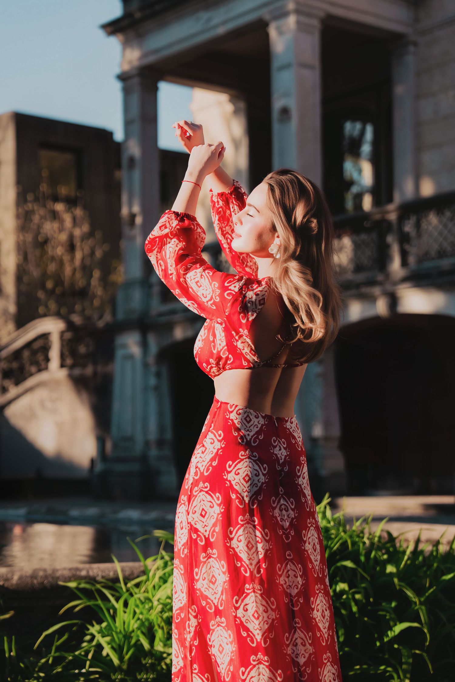 Woman in red crop top and skirt set raises arms gracefully before a classic building.