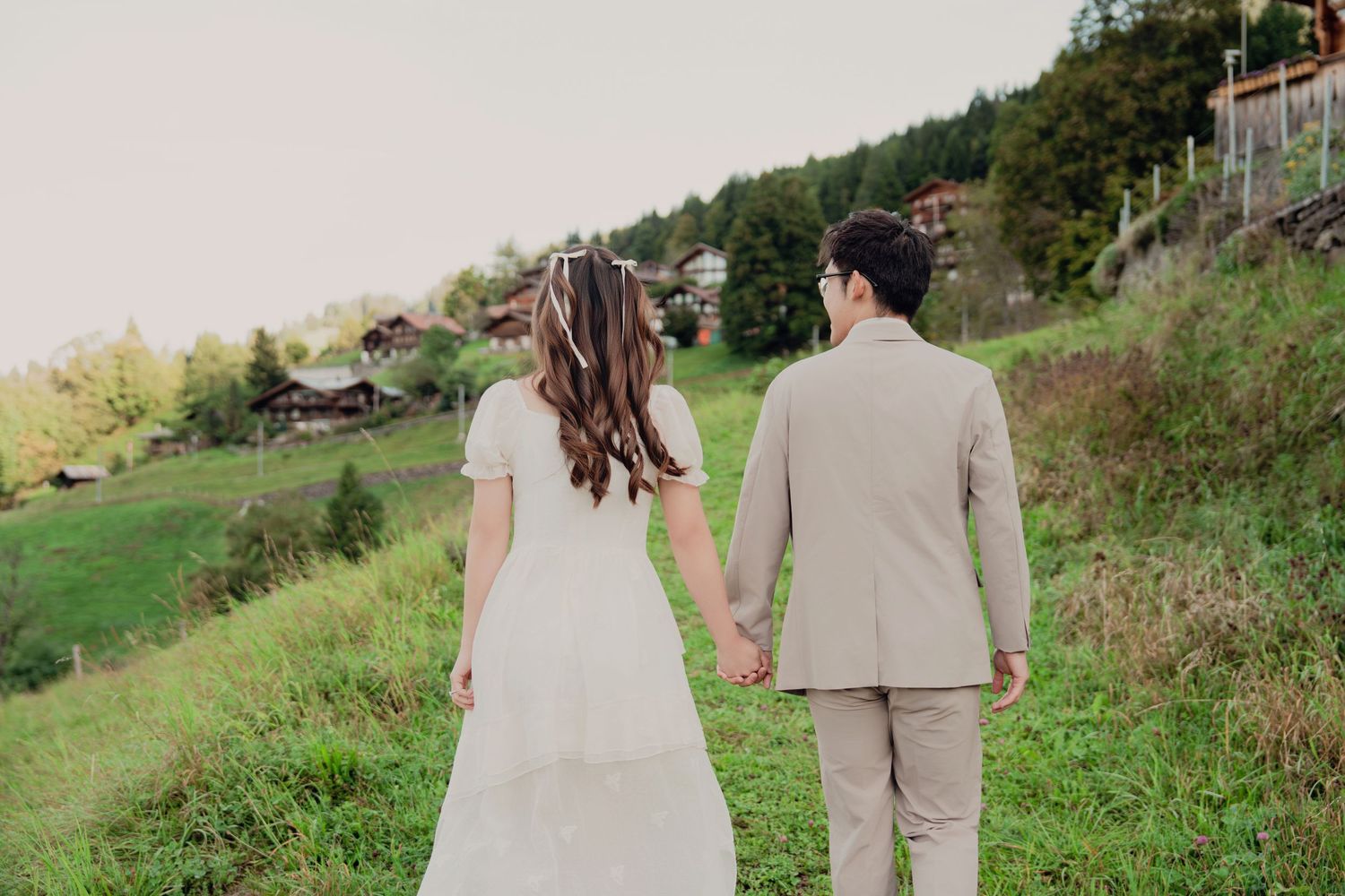 Bride in white dress and groom in beige suit holding hands walking through scenic green mountain landscape.