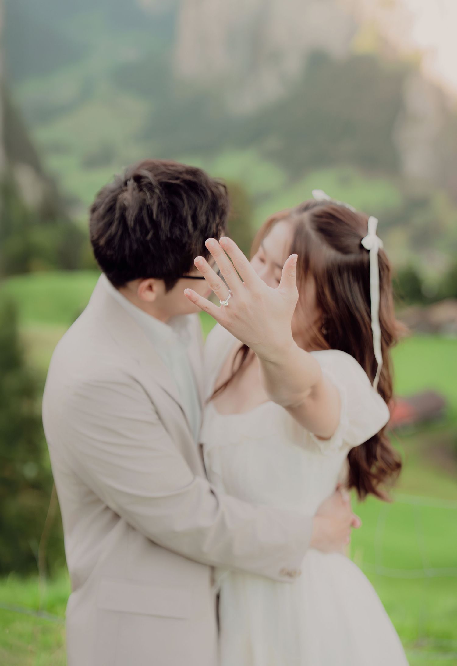 Couple sharing a kiss while showing off an engagement ring with a lush green Alpine landscape in the background.