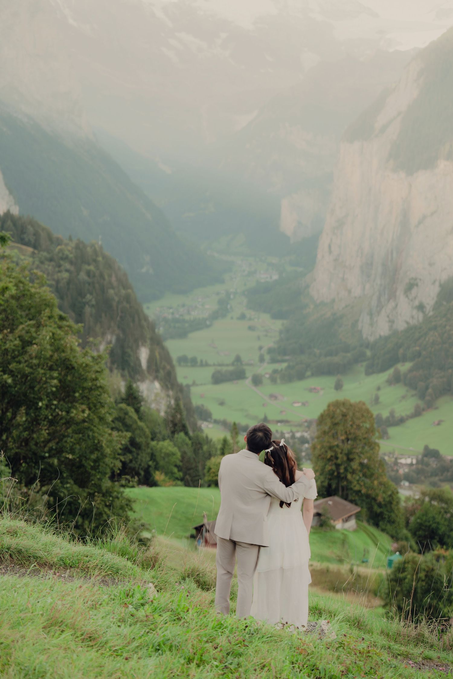 Couple standing together on a misty Alpine hillside overlooking a lush green Swiss valley with dramatic cliffs.