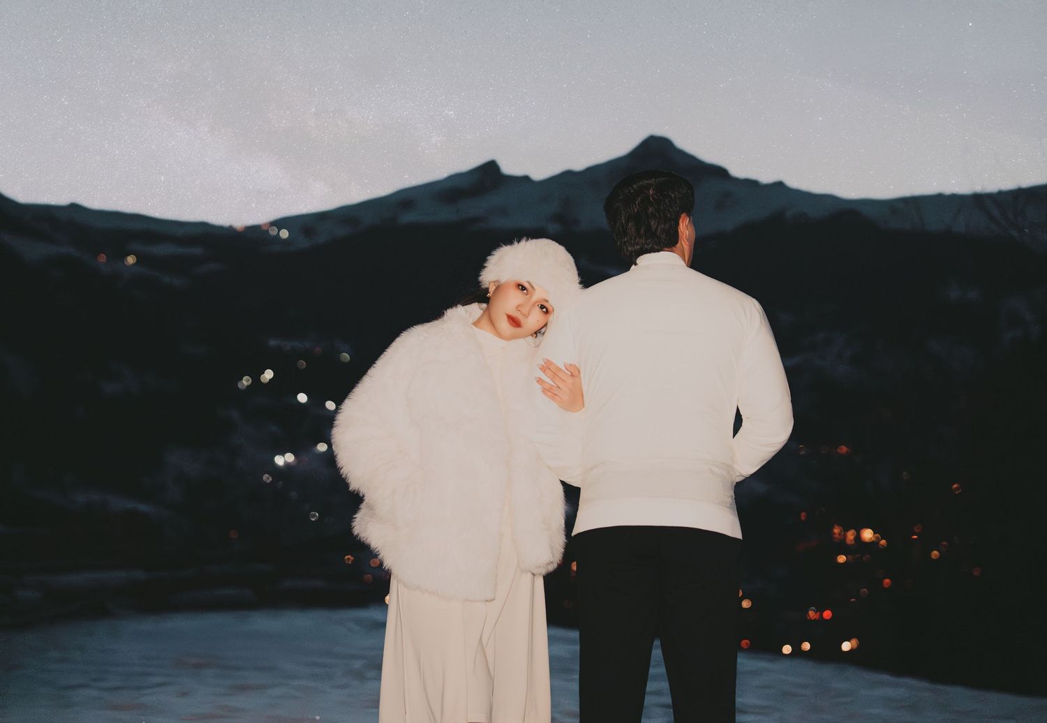 Couple in white winter outfits standing together overlooking a mountain village illuminated at dusk.