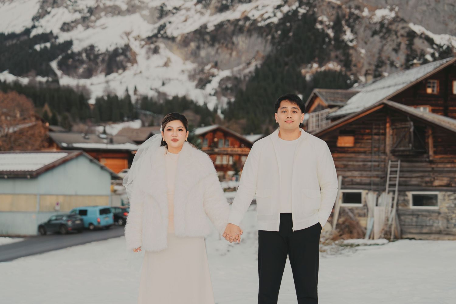 Couple holding hands in matching white outfits standing in snow with Swiss alpine chalets behind them.