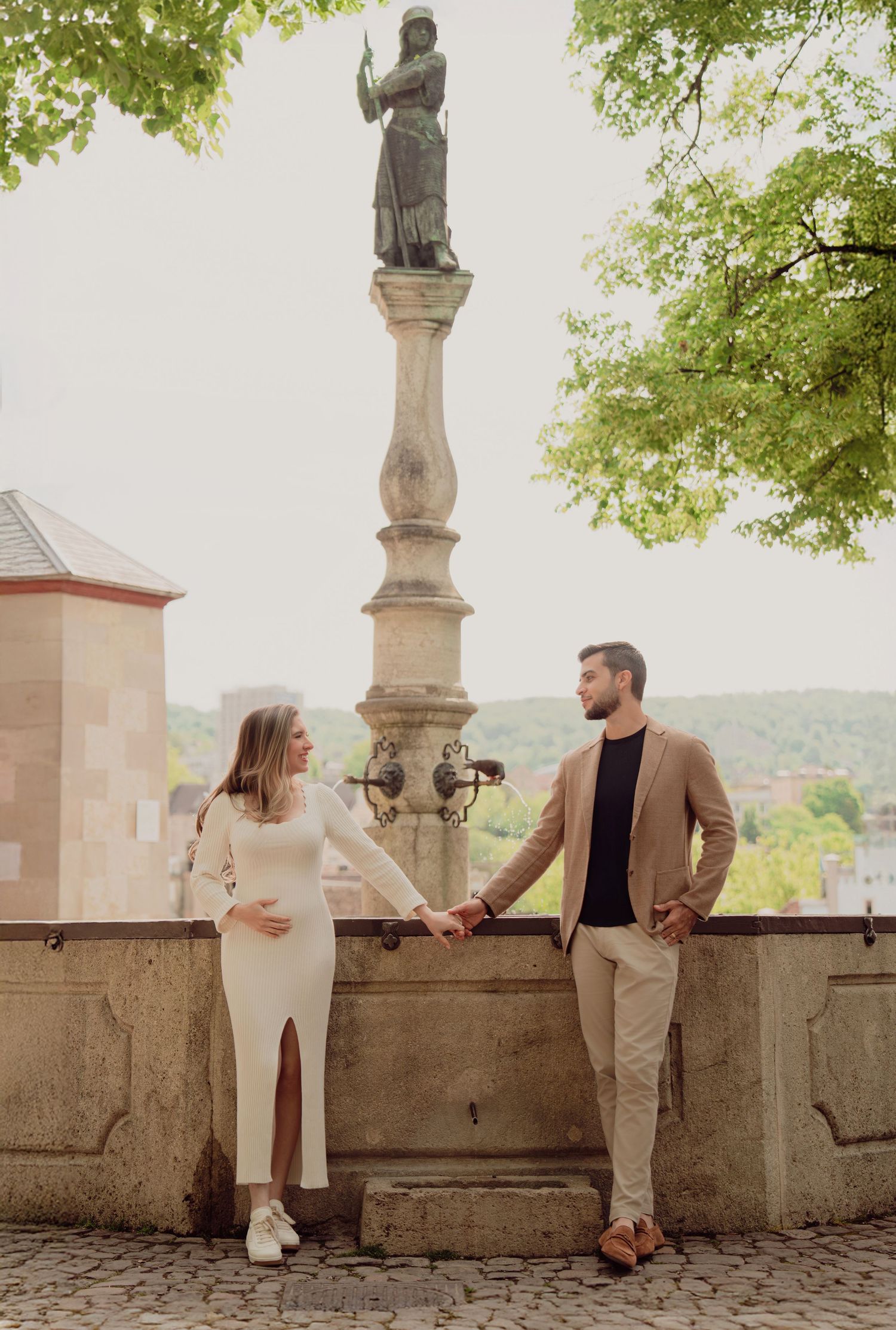 Expecting couple holding hands beside a historic stone statue column, woman in white dress, man in tan blazer.
