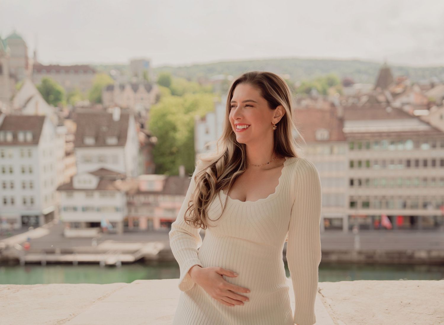 Glowing pregnant woman in cream ribbed dress cradles baby bump with Zurich city view behind her.