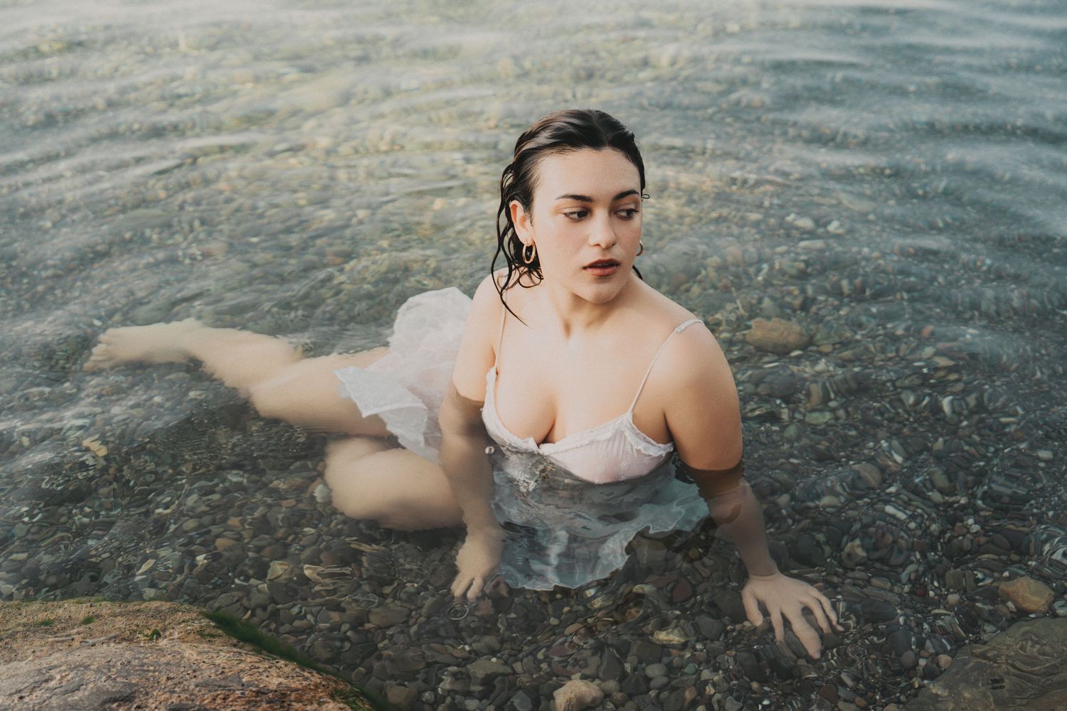 Woman in wet white dress sitting in shallow rocky water, looking upward with serene expression.