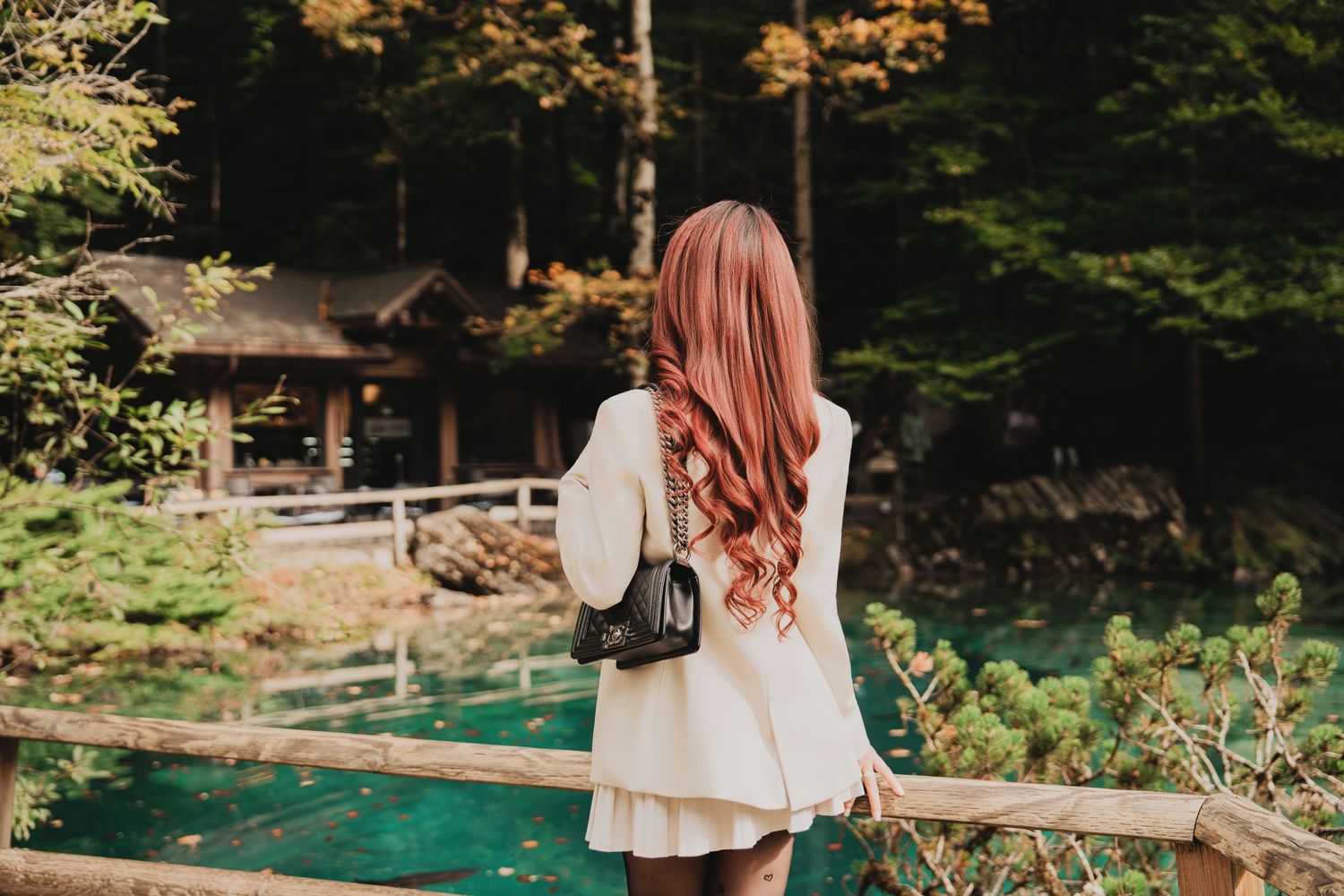 Red-haired woman with black backpack admiring turquoise alpine lake surrounded by autumn forest.