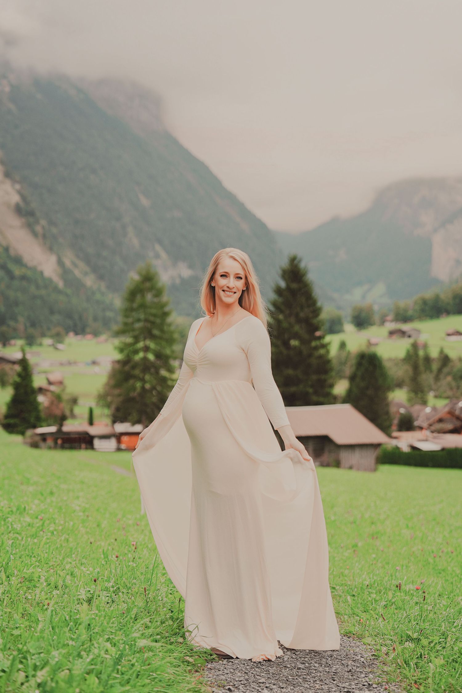 Radiant pregnant woman in long cream dress smiling while holding her gown in a picturesque Swiss mountain setting.