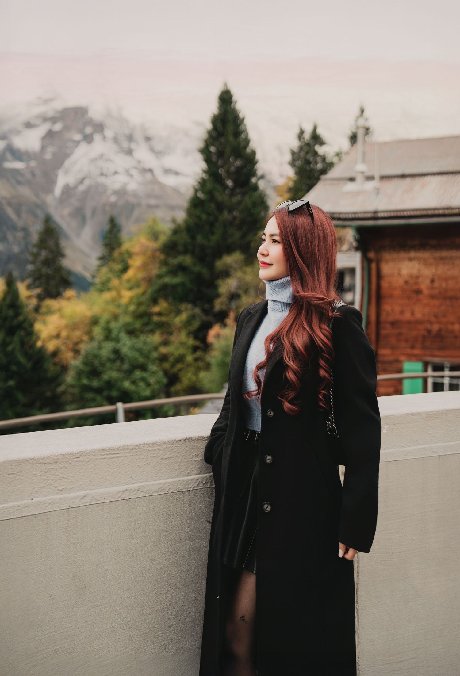 Woman in black maxi coat posing on terrace with snow-capped mountains and pine trees in background.