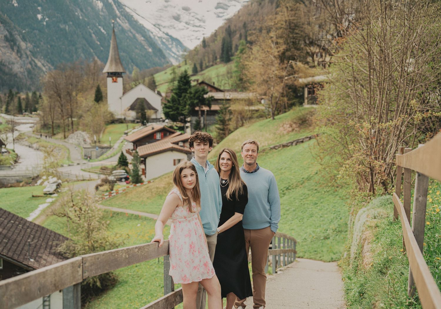 Family of four on a wooden bridge with a charming Swiss Alpine village and church below.