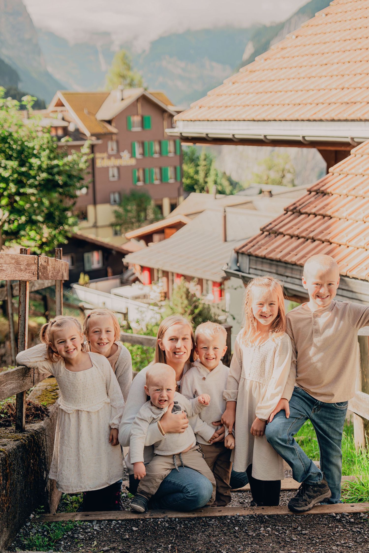 Mother crouching with six blonde children on steps with Swiss Alpine village in the background.