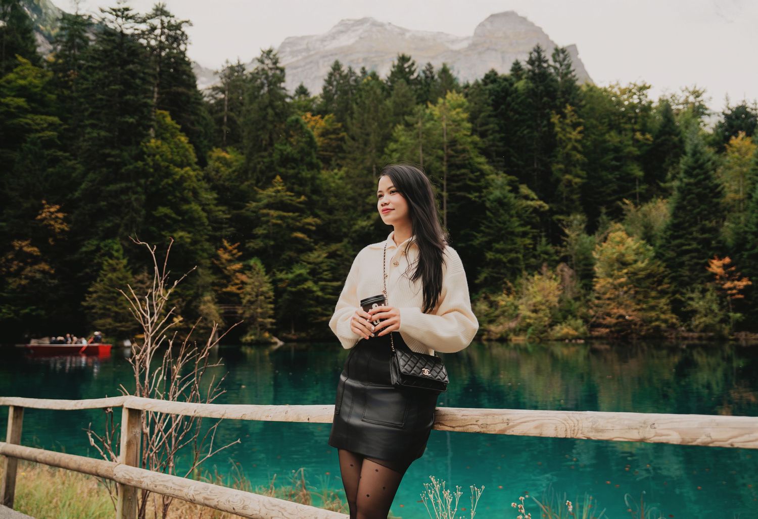 Woman in beige sweater and black leather skirt holding coffee by wooden fence at turquoise mountain lake.