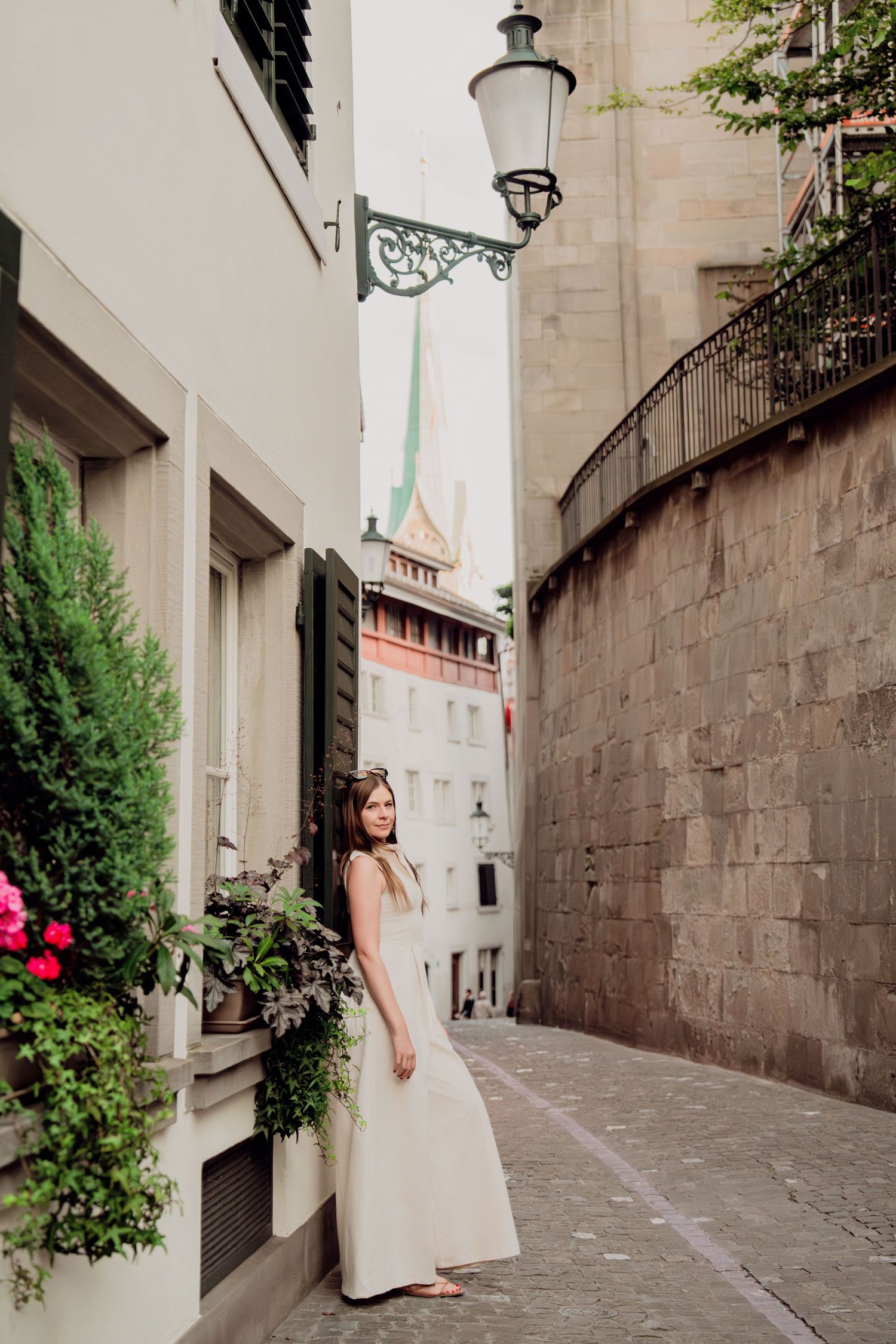 Woman in white maxi dress poses in charming narrow alley of Zurich old town with church spire.
