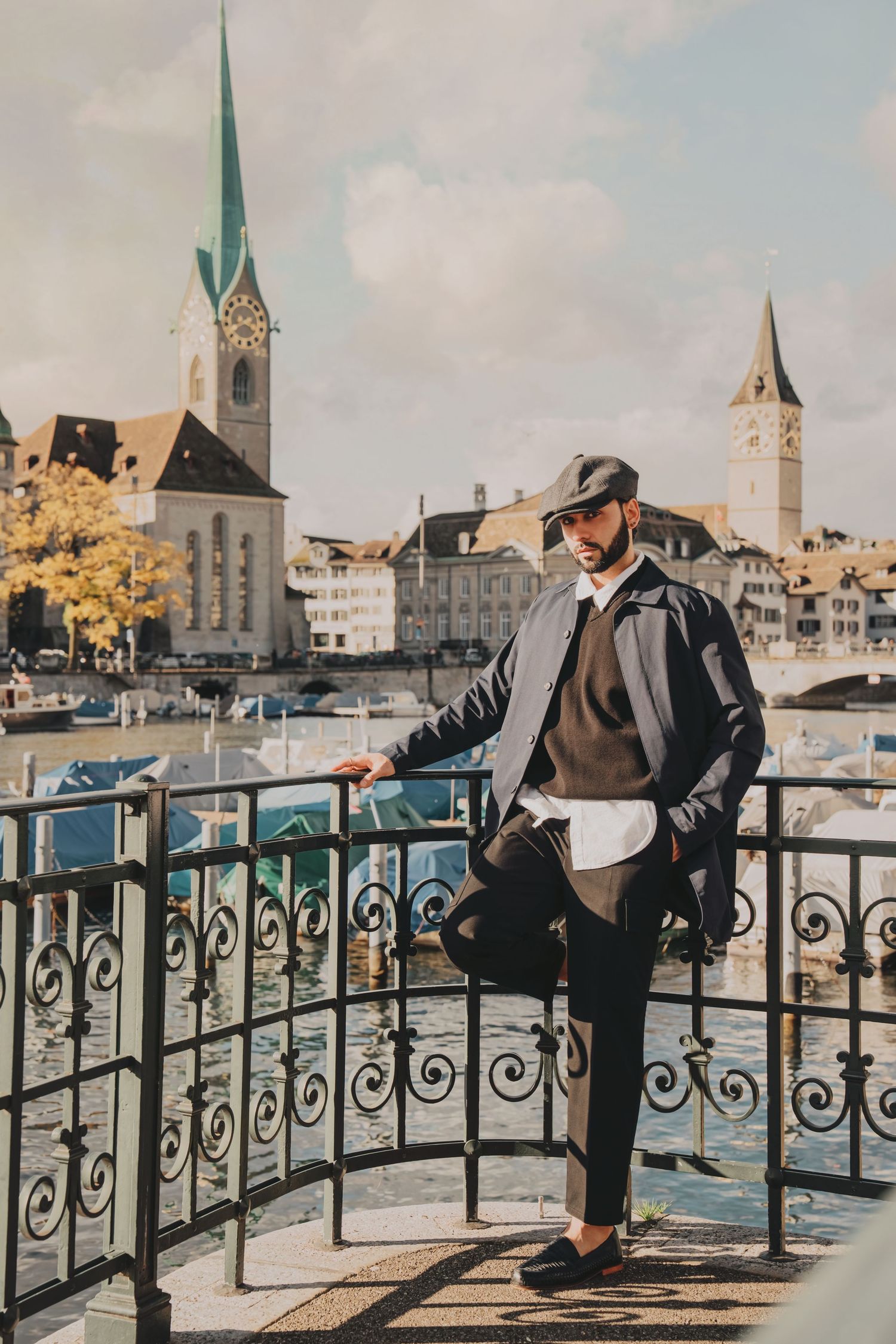 Dapper man in layered outfit and flat cap leans on ornate iron fence by Zurich river with twin church spires.