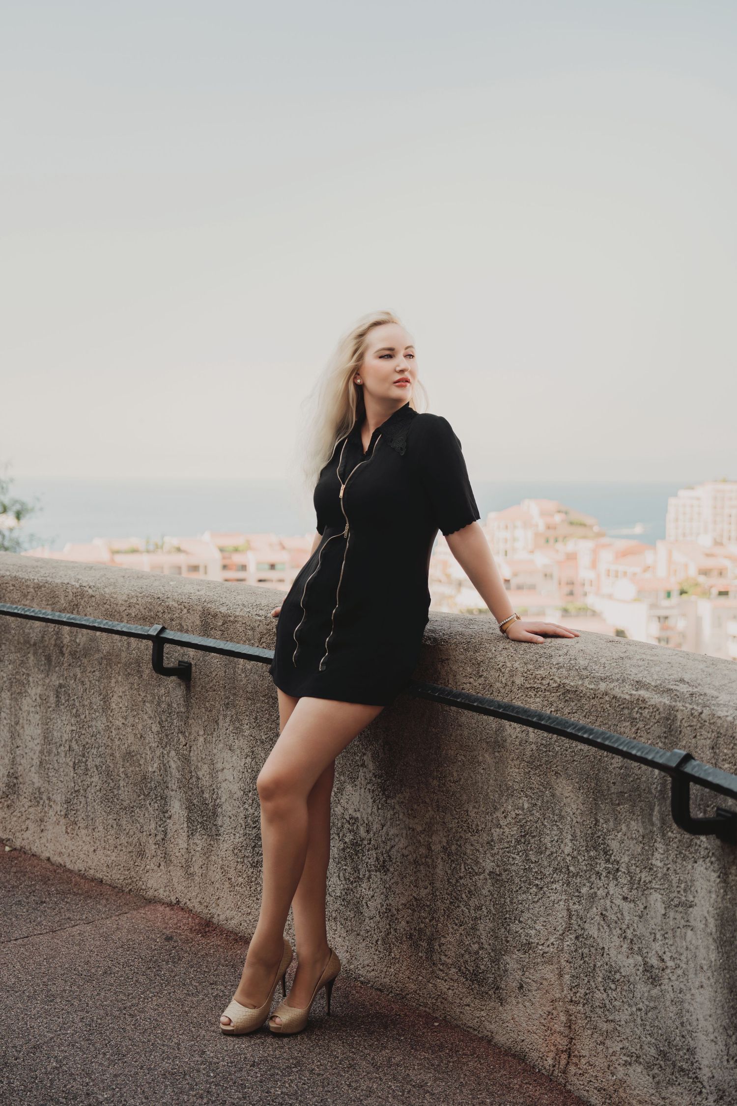 Blonde woman in black mini dress leaning on stone balustrade overlooking Monaco cityscape.