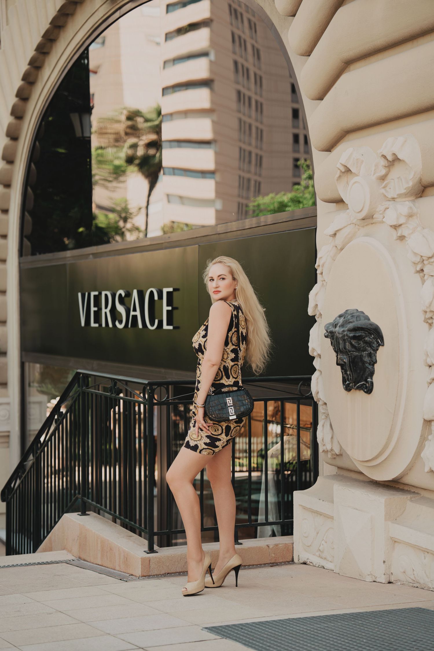 Stylish blonde woman in Versace mini dress posing outside Versace store entrance in heels.