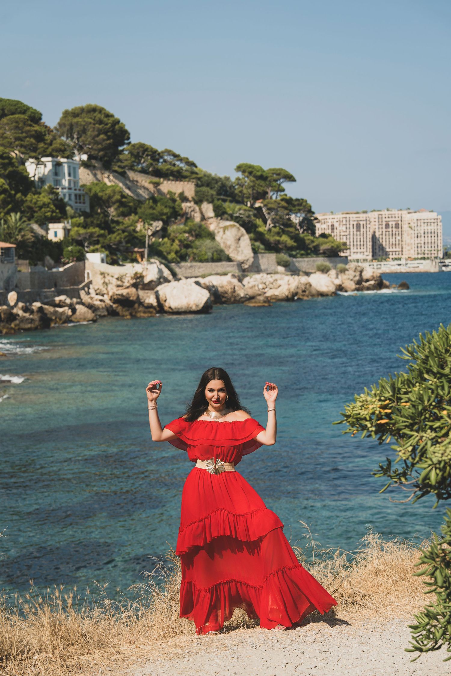 Woman in red off-shoulder tiered maxi dress posing by Mediterranean coastal scenery with turquoise water.