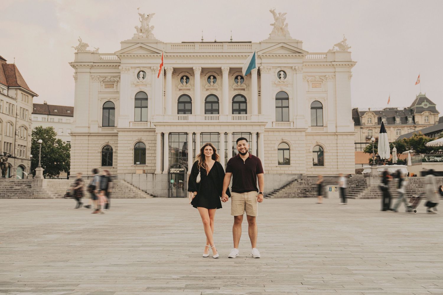 Couple in matching black and tan outfits standing before a grand neoclassical building in a European plaza.