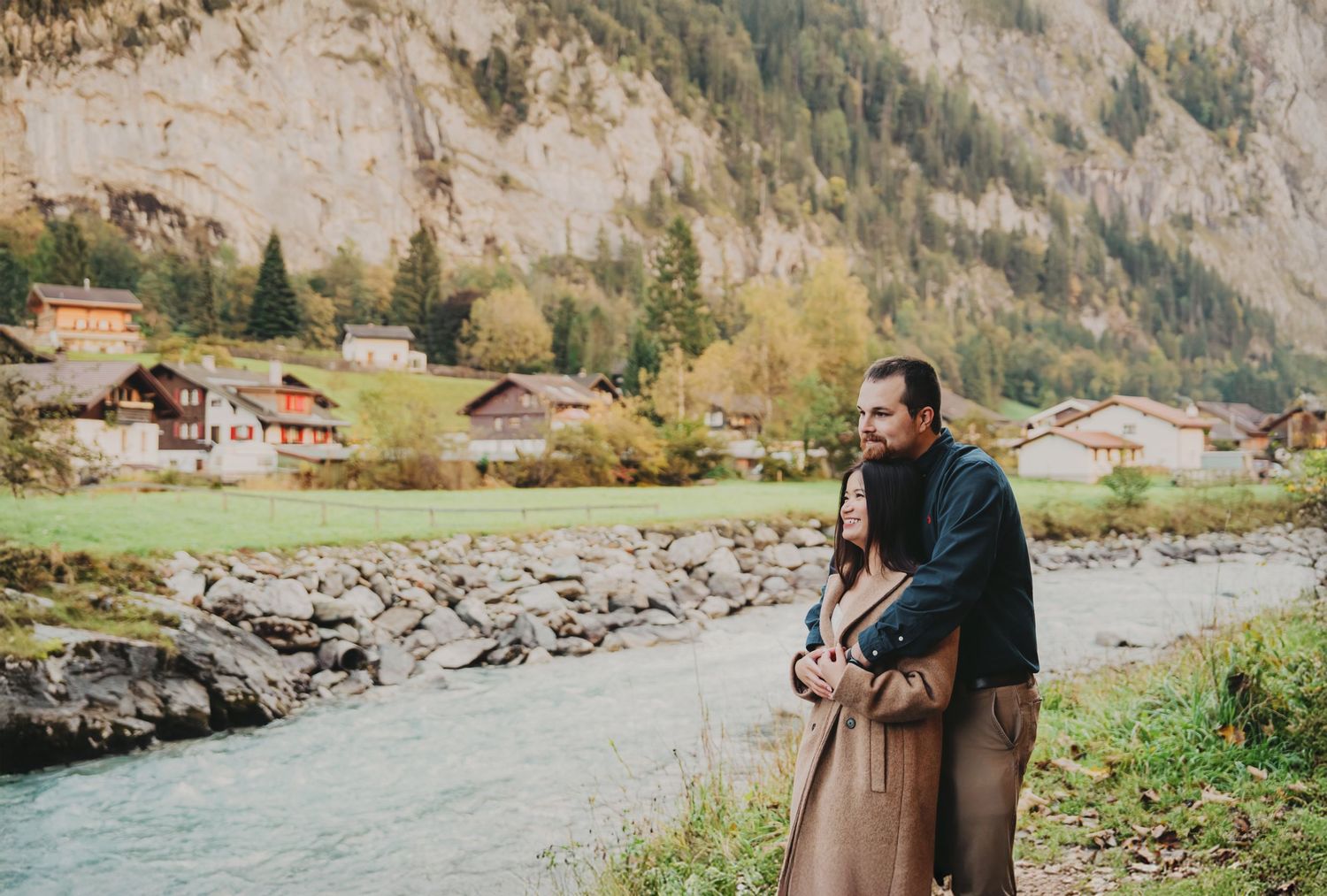 Couple embracing by a turquoise mountain river with Swiss chalets and cliffs behind.