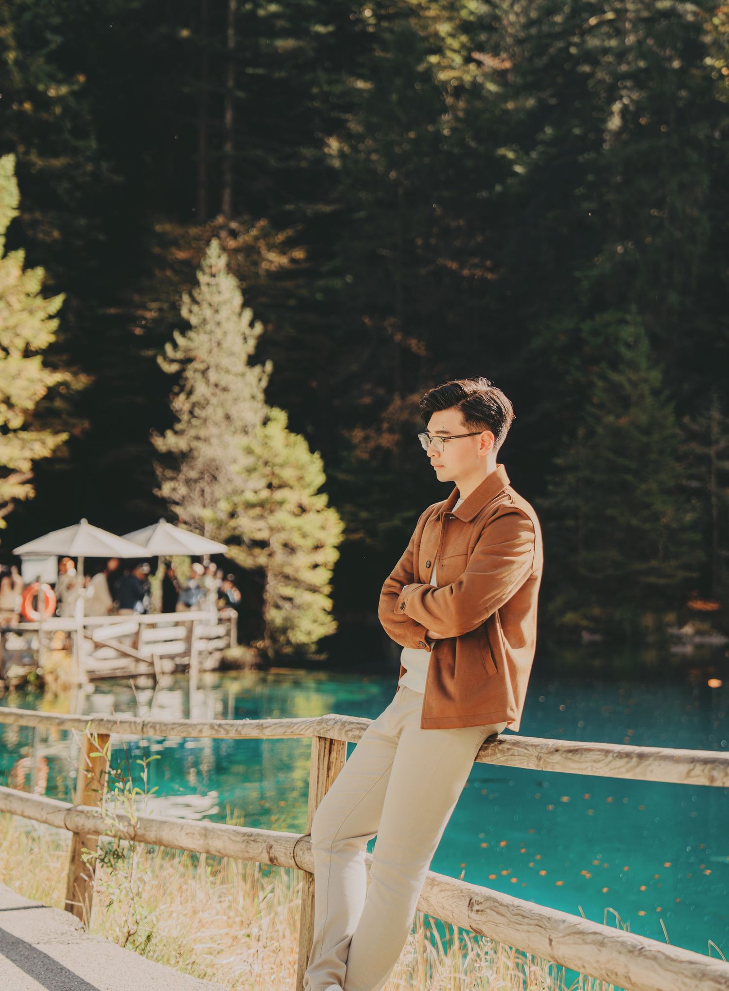 Young man in brown jacket leaning on wooden fence by turquoise lake in forest.