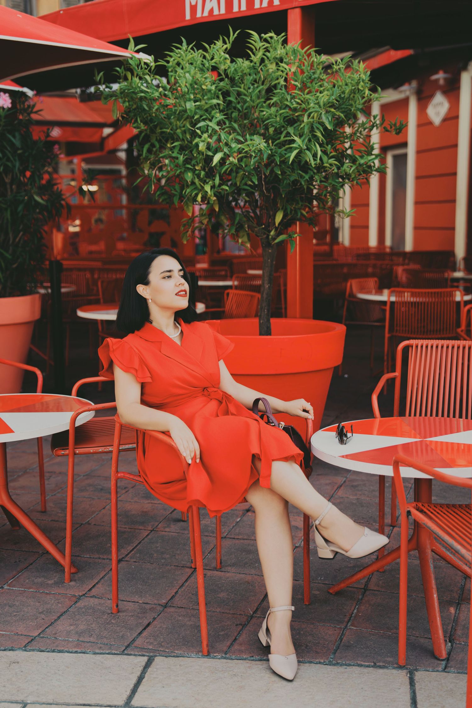 Stylish woman in red wrap dress sitting at outdoor cafe terrace surrounded by matching red chairs and decor.