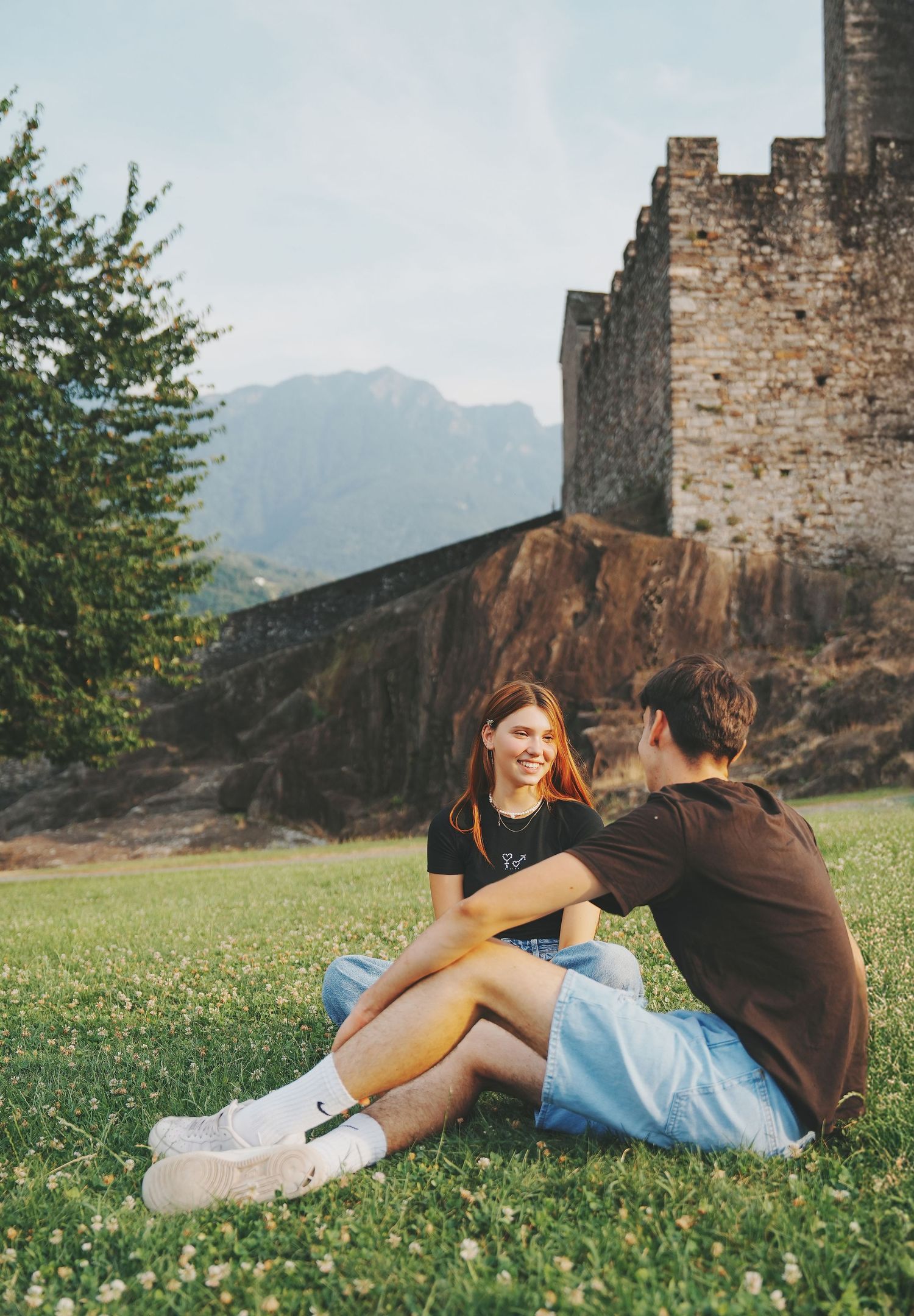 A young couple sitting on a grassy lawn beside an ancient stone castle wall with mountain views.