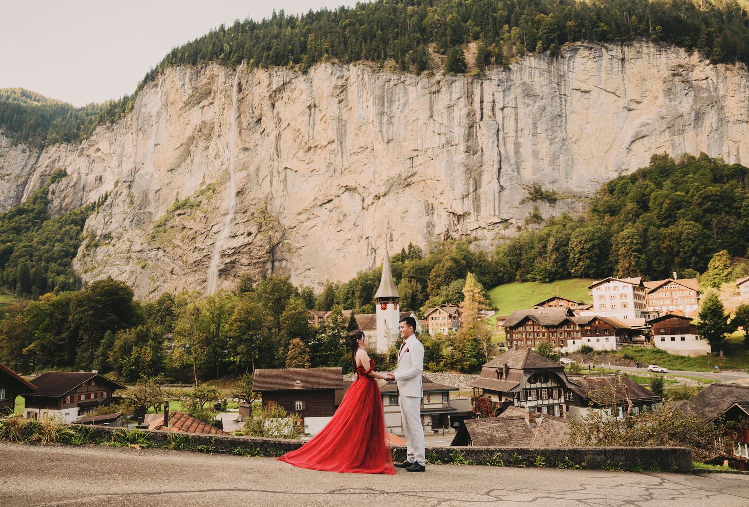 Couple standing together with dramatic Swiss cliffs and village church visible in the background.