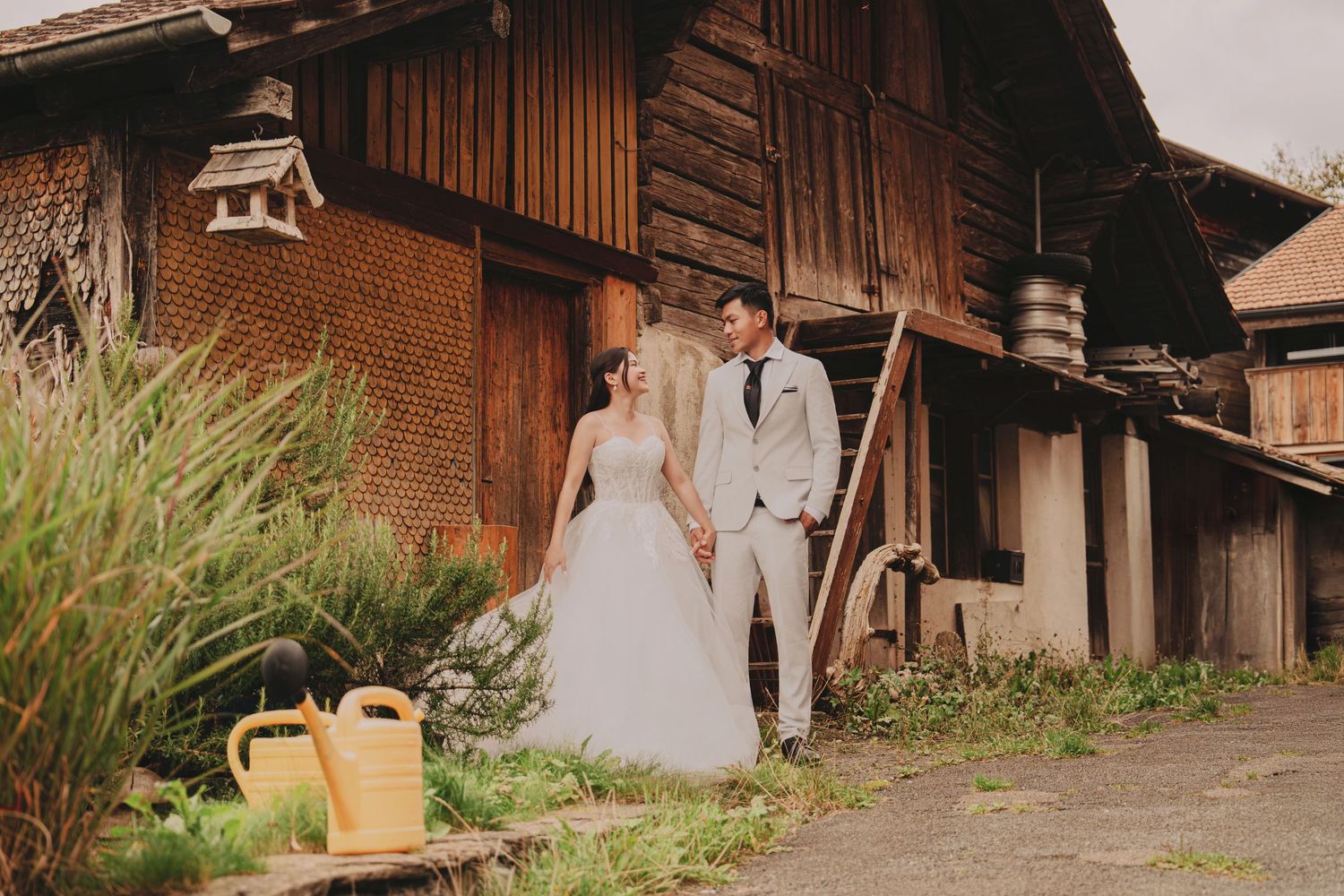Bride and groom holding hands near rustic wooden barn in picturesque Swiss countryside setting.