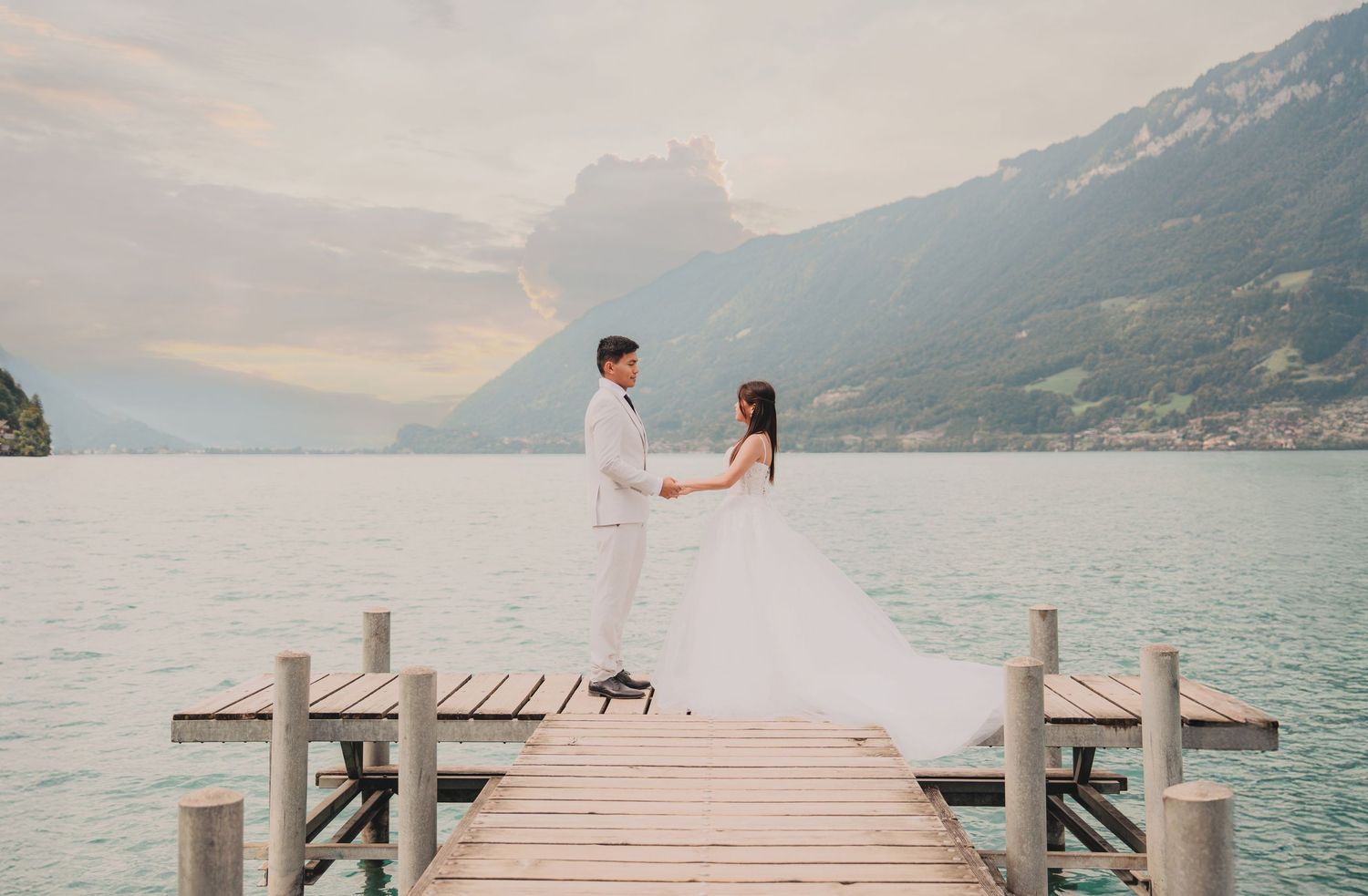 Bride and groom face each other holding hands on a wooden dock with stunning alpine lake and mountains.