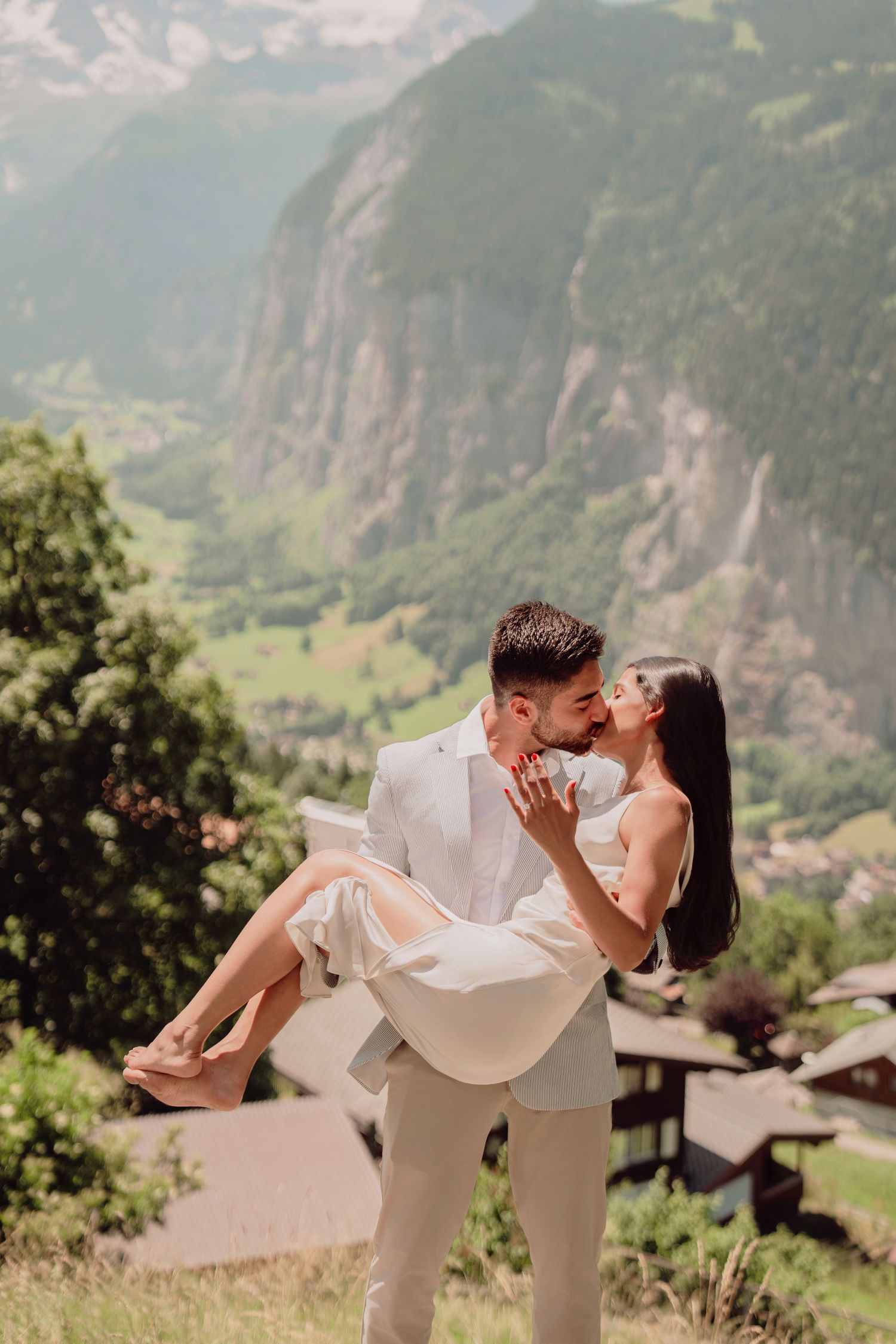 Groom lifting and kissing bride against stunning Swiss Alps valley and waterfall backdrop.