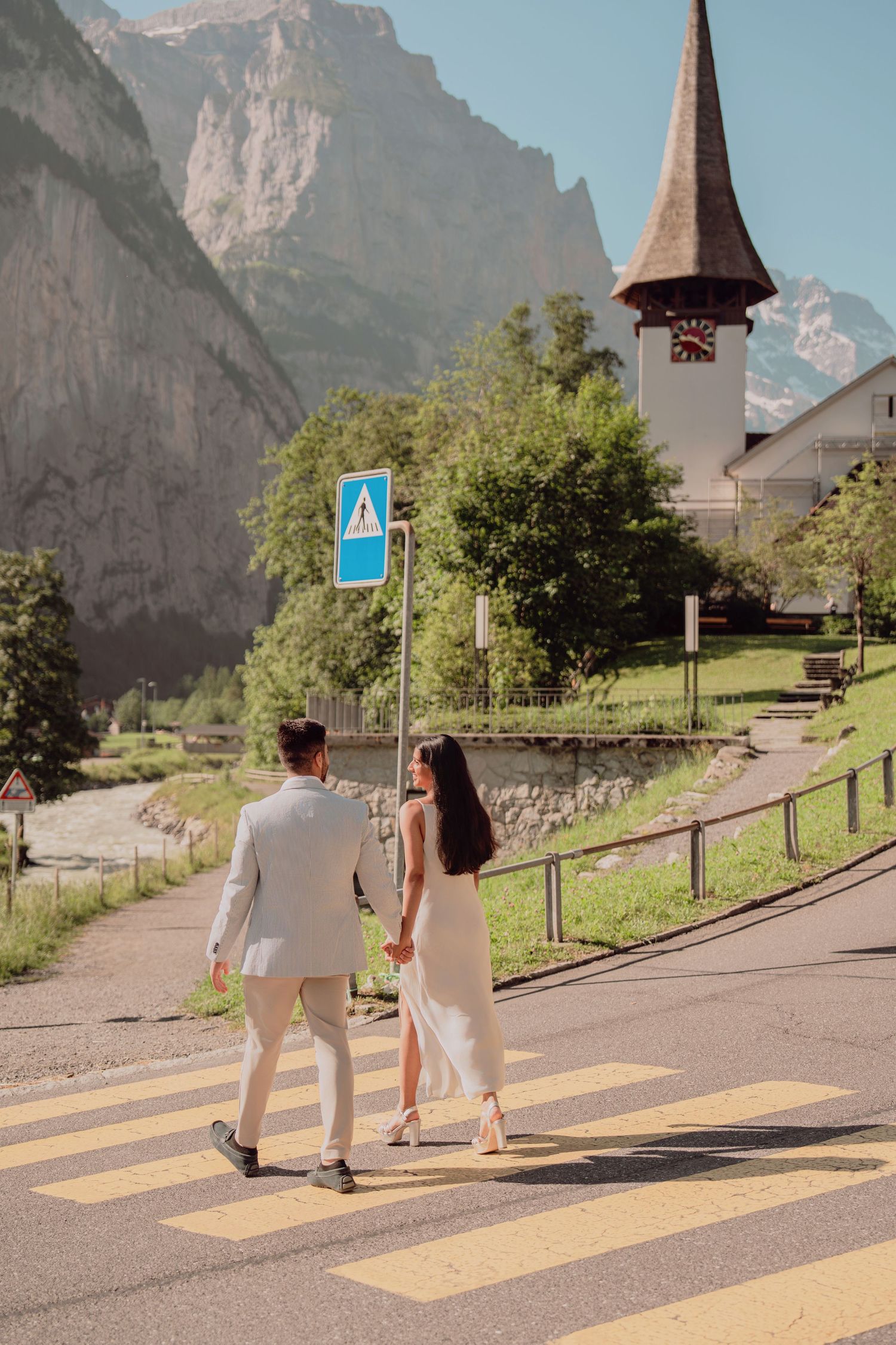 Couple walking hand-in-hand across a zebra crossing with a Swiss church steeple and alpine mountains behind them.