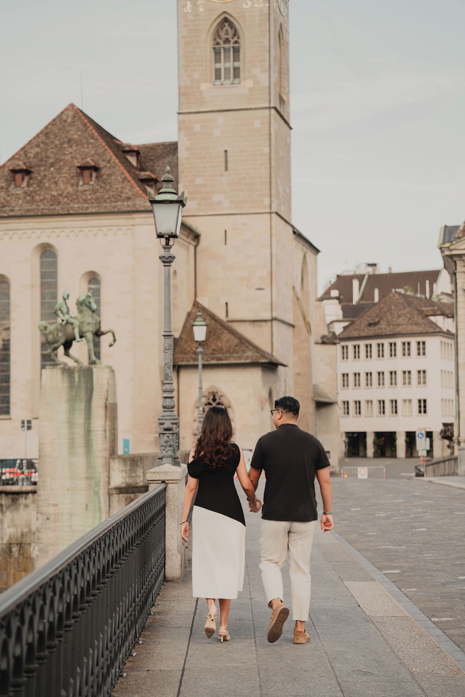 Couple walking hand in hand along a Zurich bridge with a historic church tower and old buildings.