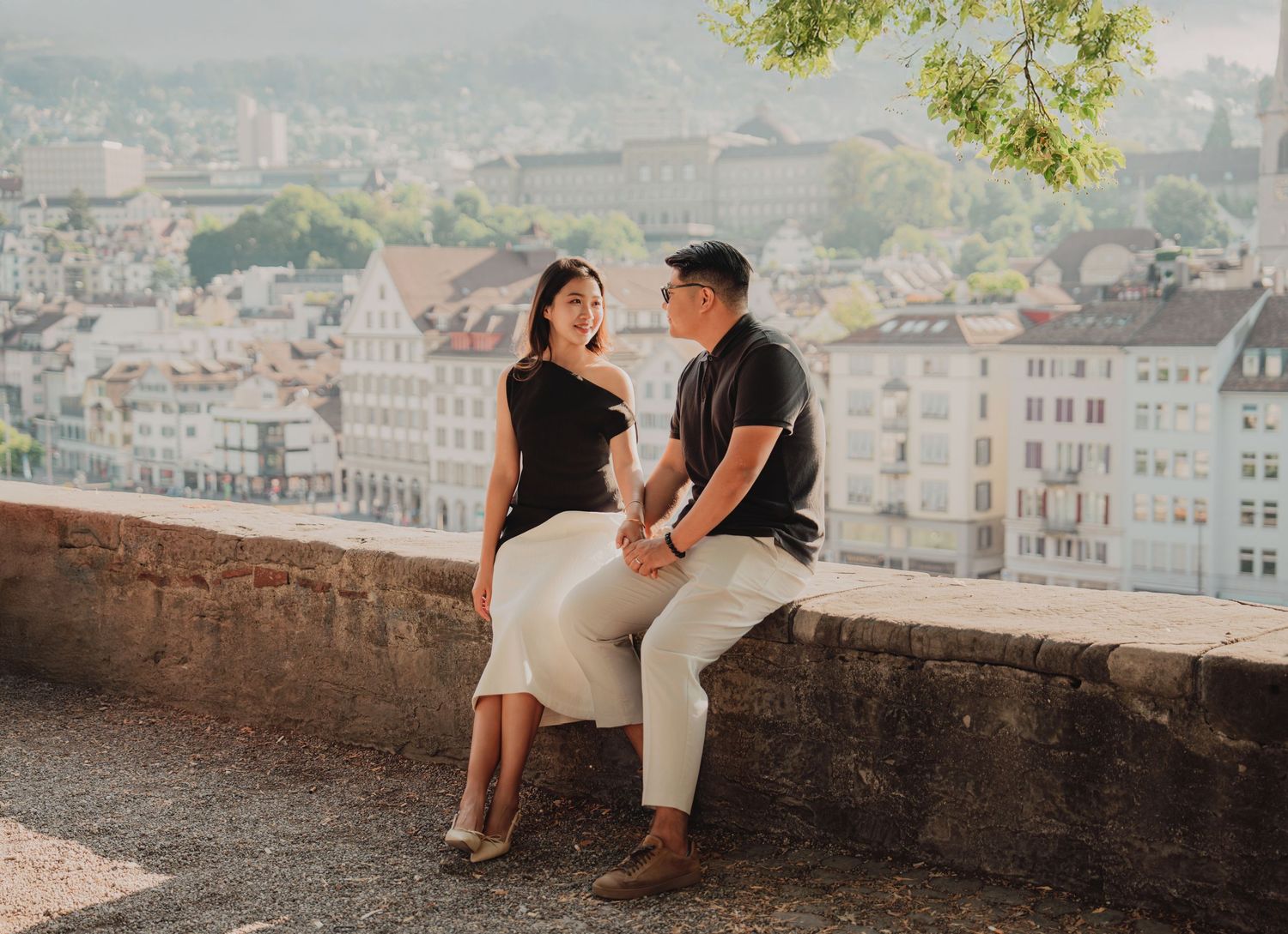 Couple sitting on a stone wall laughing together with the beautiful Zurich city panorama in the background.