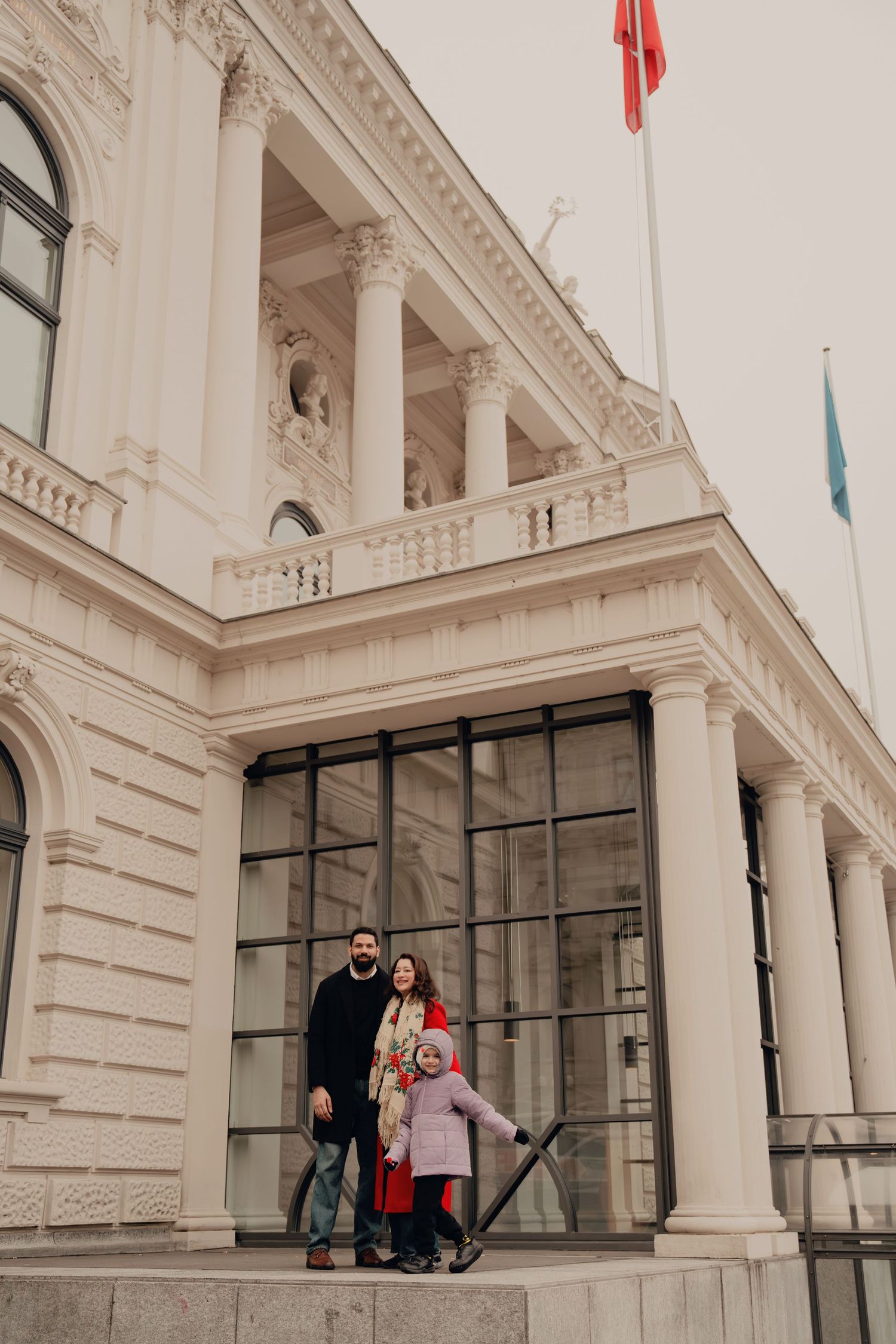 Family of three posing on steps before a grand neoclassical building with columns and flags.