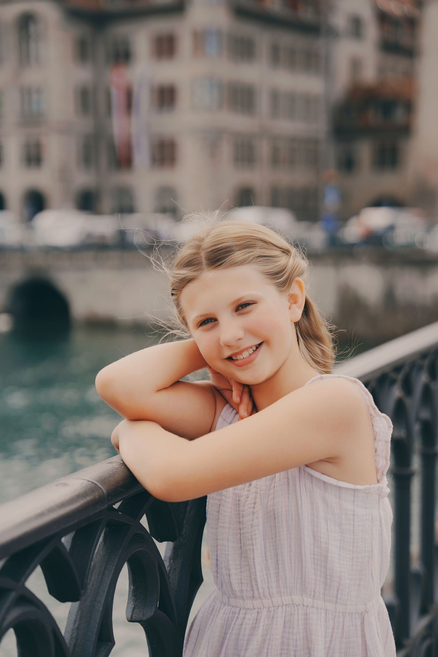 A smiling young blonde girl leans on a black iron bridge railing in Zurich, Switzerland.