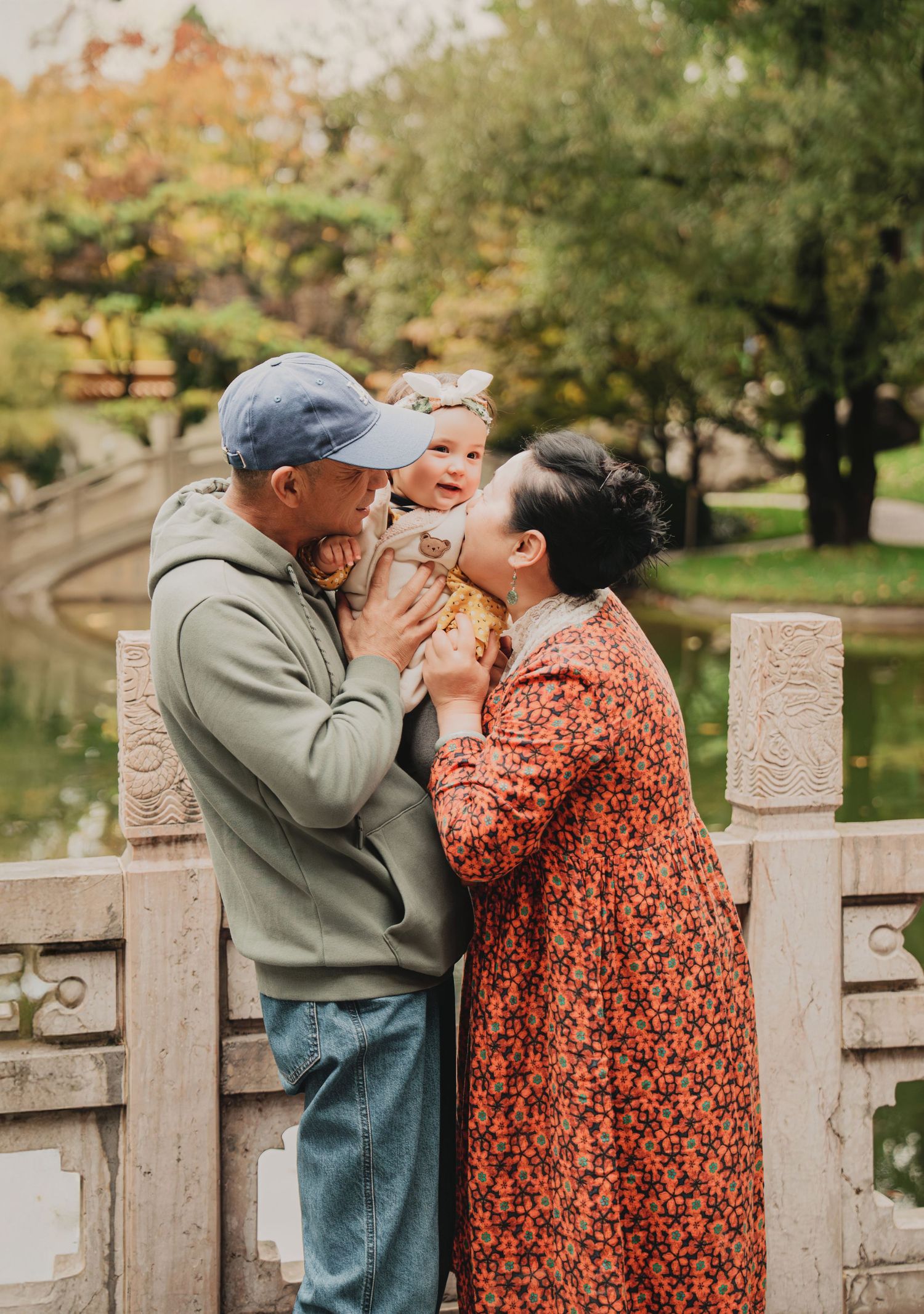 Grandparents kissing and cuddling a laughing baby girl by a serene garden pond.