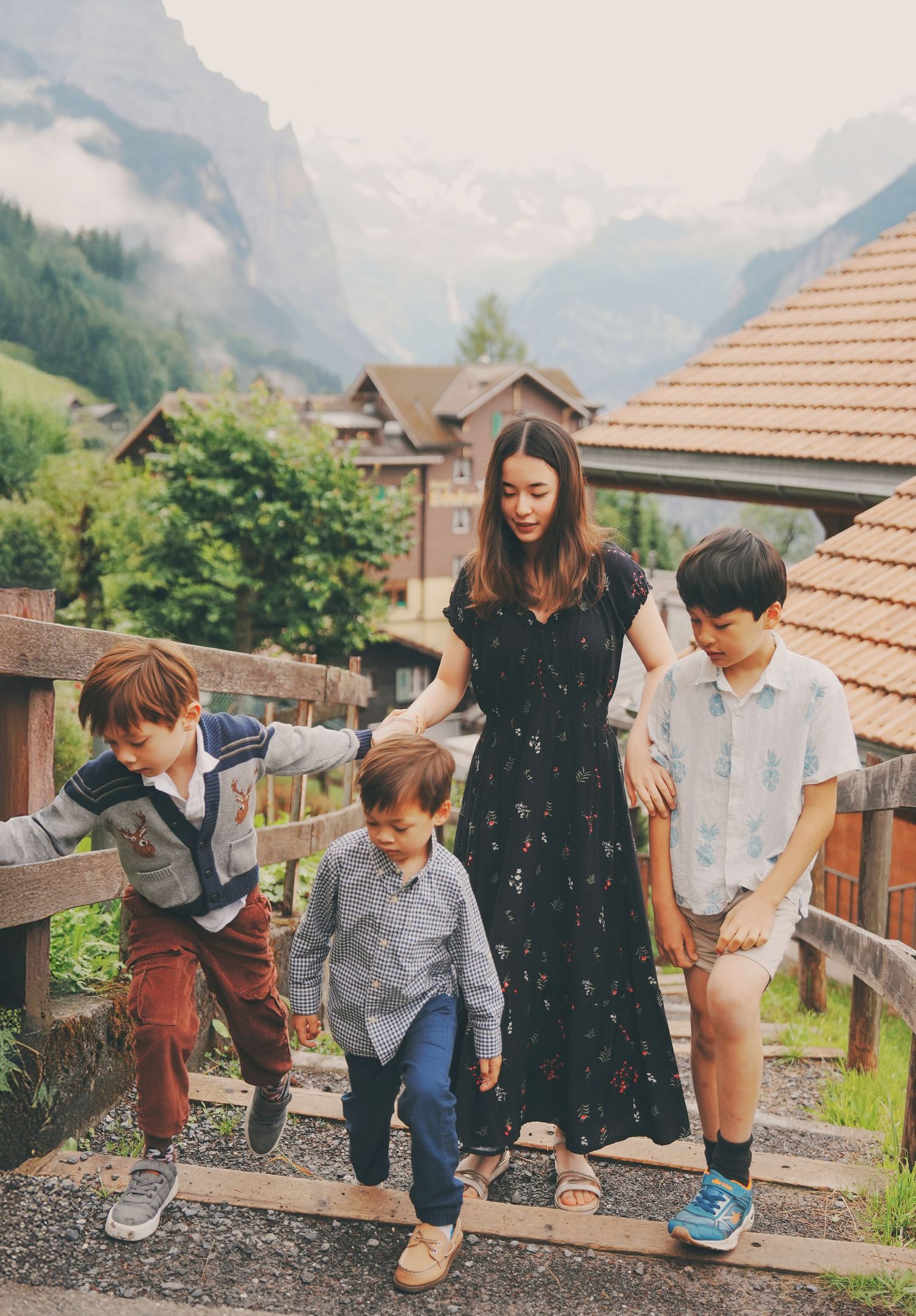 A woman in a black floral dress walks with three boys on wooden steps in a Swiss mountain village.