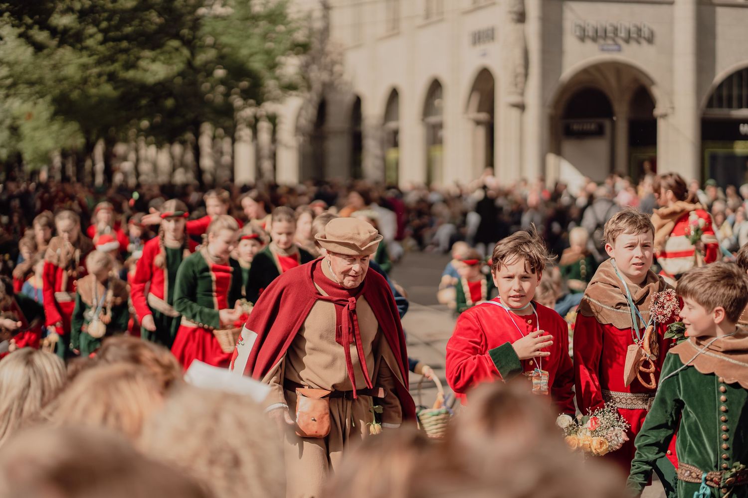 Participants in red and brown historical costumes march through a crowded city street during a festive parade.