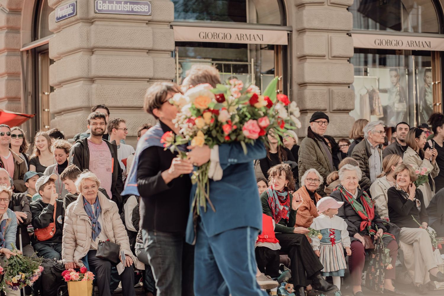 Man in blue suit presenting a large bouquet of pink and white flowers to someone in a crowd.
